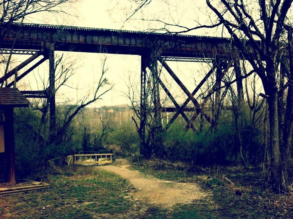 A weathered railway trestle bridge stands tall above a dirt path, surrounded by bare trees and underbrush. The scene depicts a blend of nature and industrial architecture, with a wooden sign structure visible on the left side of the image. Soft lighting creates a serene atmosphere. Devou Park mountain bike trail.