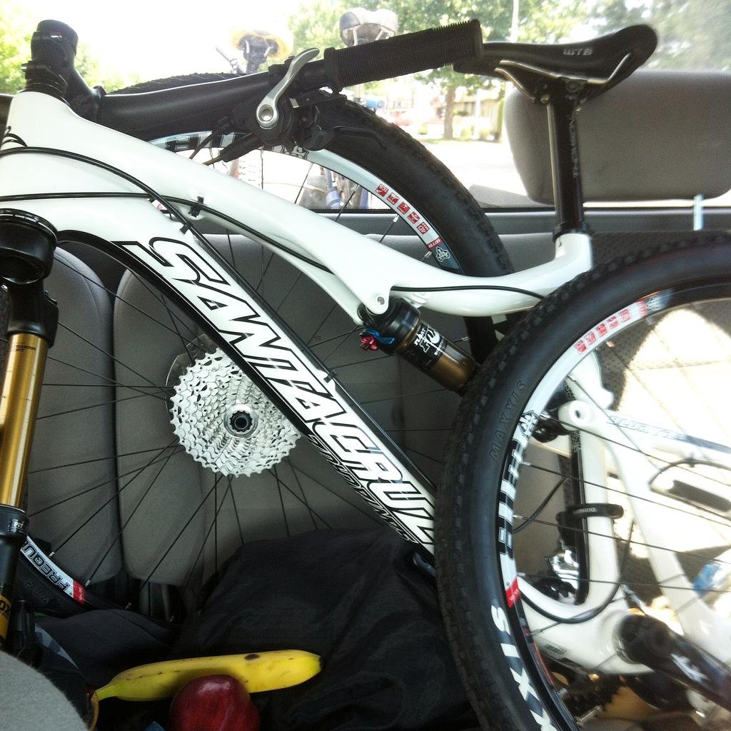 Santa Cruz Blur: A mountain bike with a white frame is positioned in the back seat of a car, partially obscured by the seat. Also visible are a bunch of bananas and an apple resting on a dark cloth. The interior of the car is softly lit, and parts of the bike, including the wheels and gear cassette, are clearly visible.