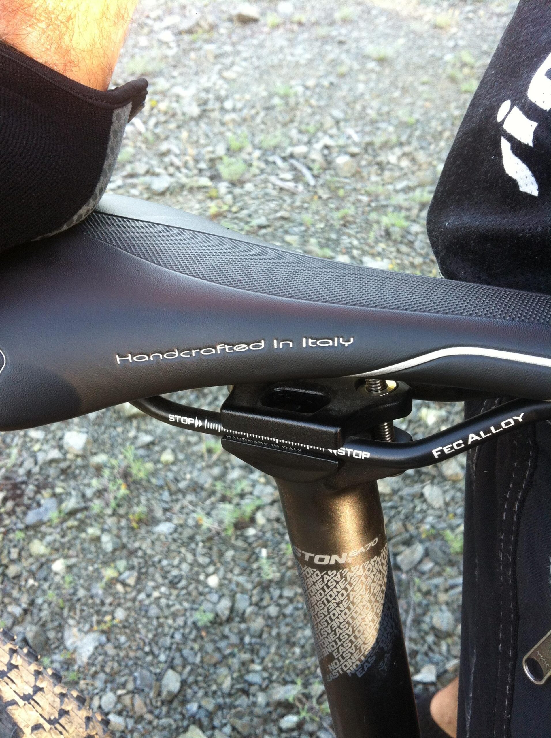Intense 2014 Spider 29c: Close-up of a bicycle saddle with the text "Handcrafted in Italy" visible. The image shows a portion of the seat post and a hand resting on the saddle, with a background of gravel and grass. The seat post is marked with the brand name "Easton" and includes details on the adjuster.