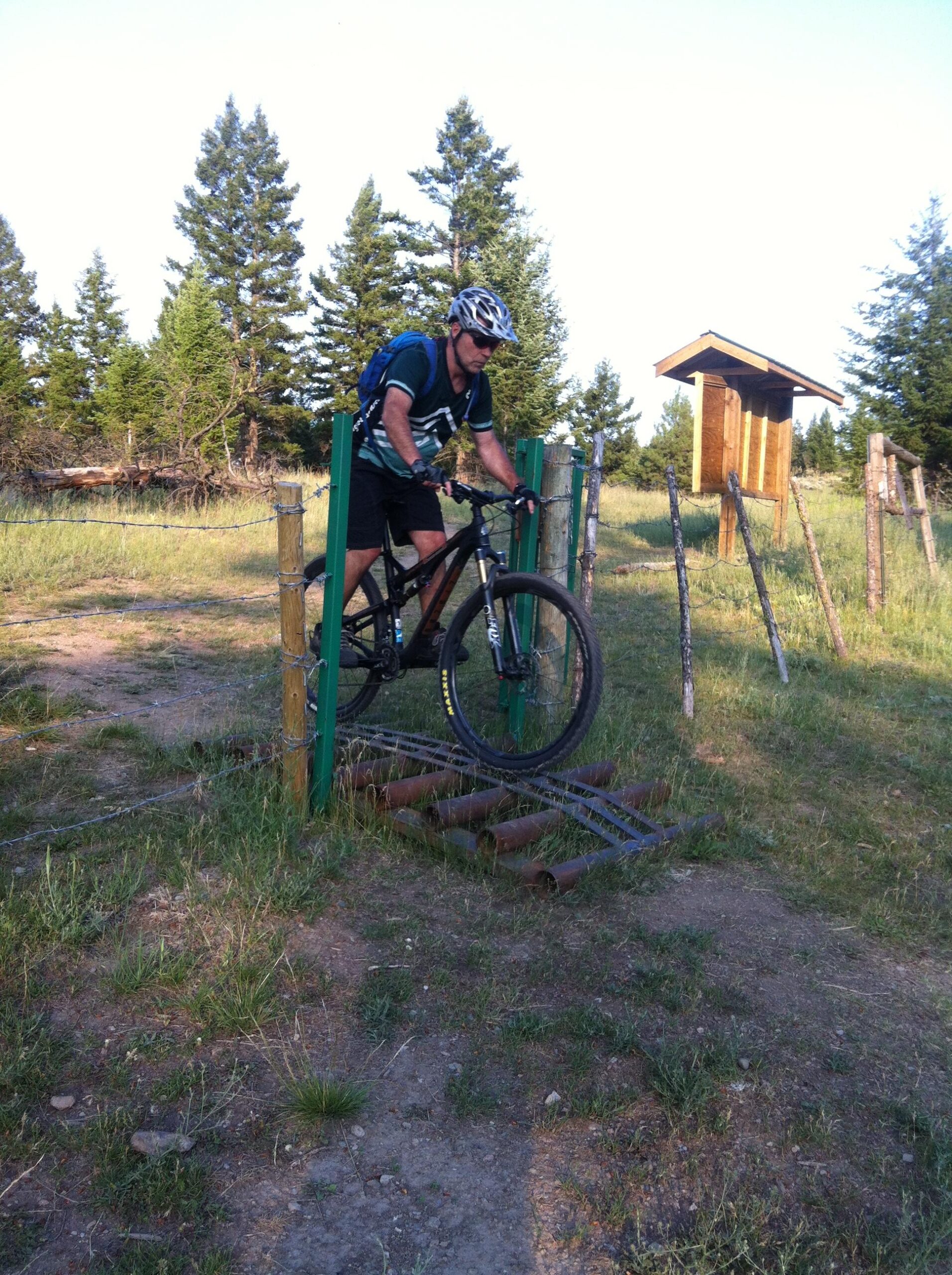 A cyclist wearing a helmet and a backpack skillfully navigates over a log barrier on a mountain bike trail, surrounded by grassy fields and tall trees under a clear sky. A wooden structure is visible in the background. Pineview mountain bike trail.