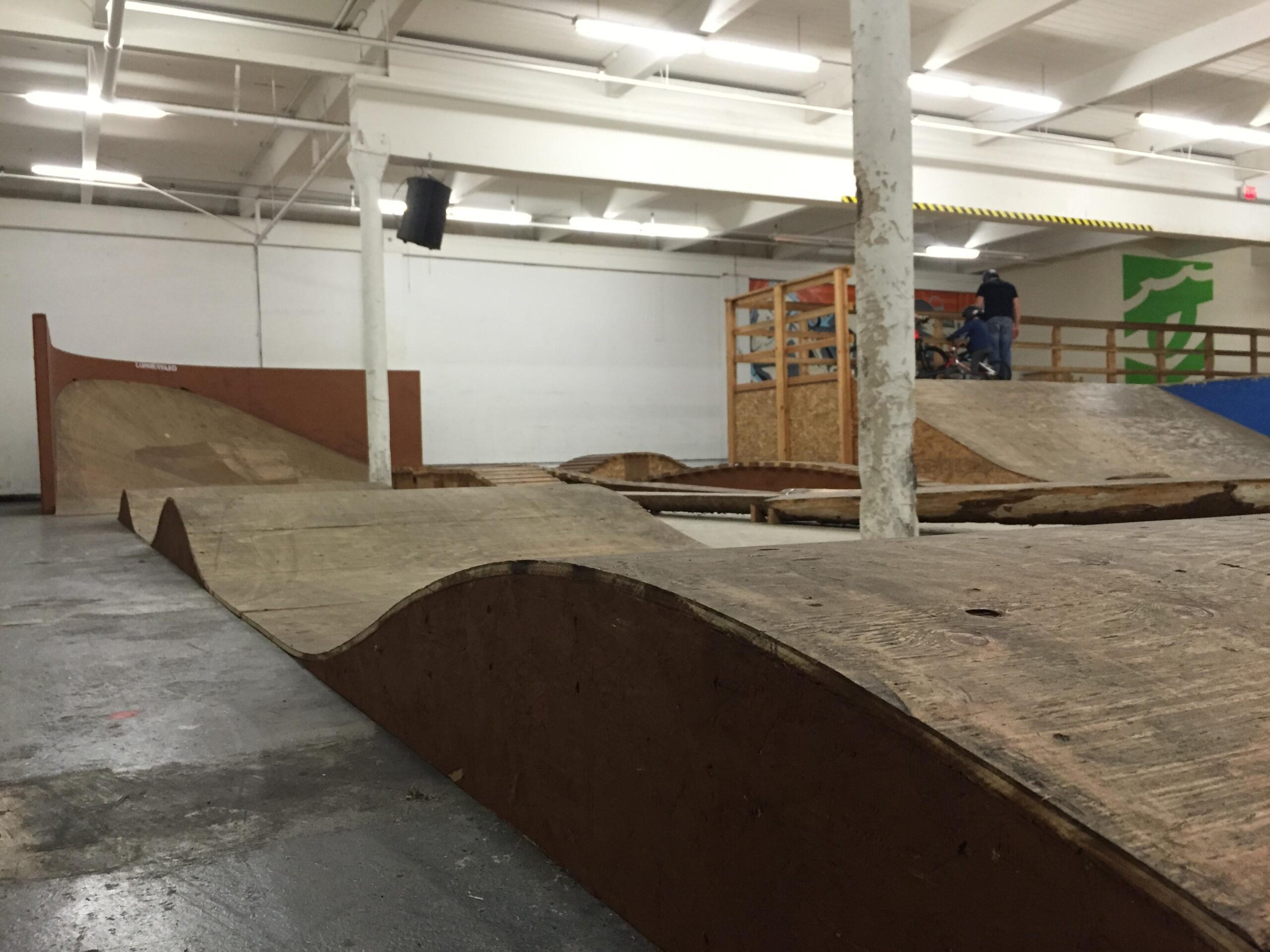 Indoor skatepark featuring wooden ramps and jumps, with a rider in the background preparing on a bike. The space has a low ceiling and ample lighting, creating a functional atmosphere for BMX or skateboarding activities. Lumberyard Indoor Bike Park mountain bike trail.
