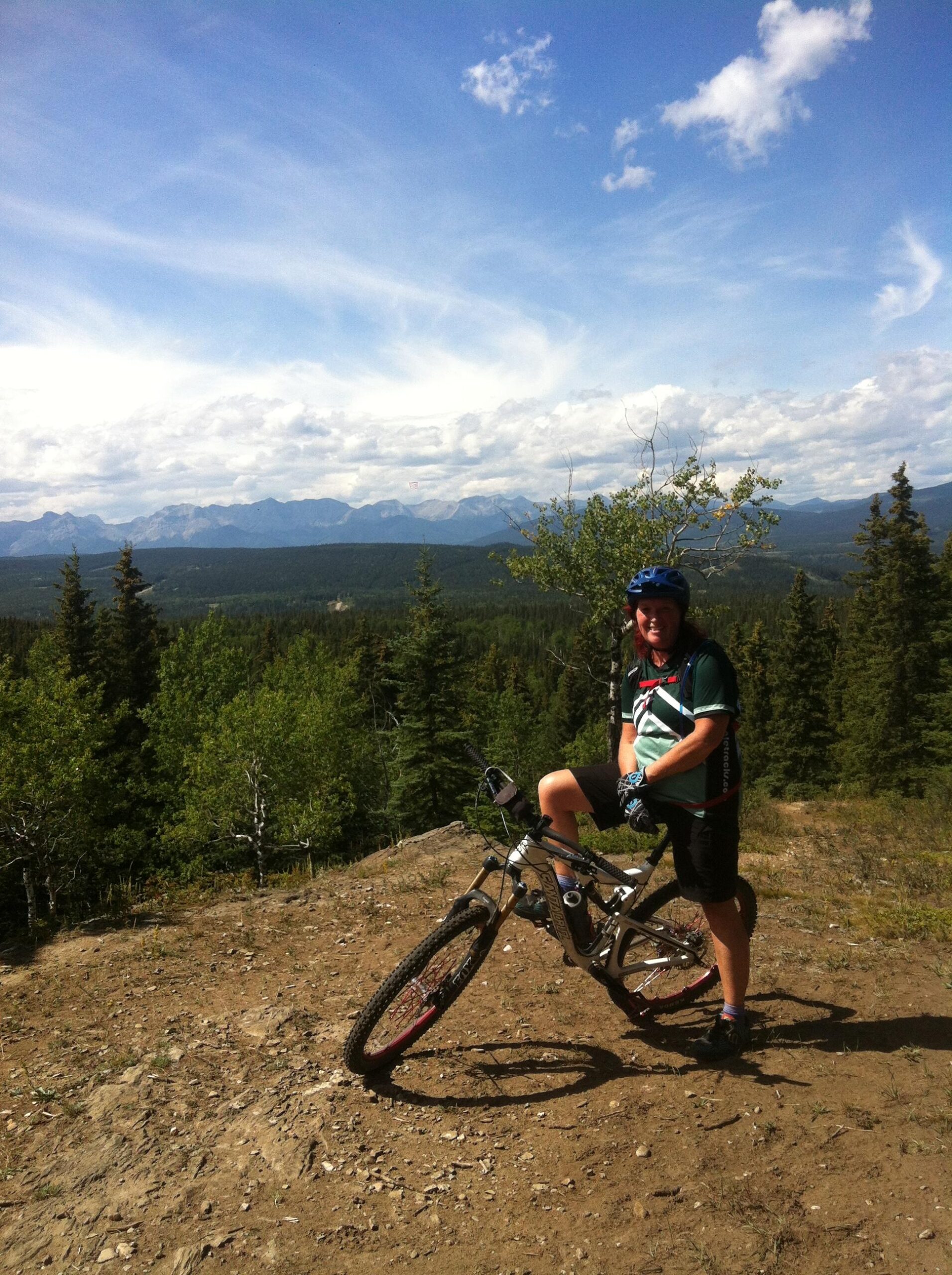 A mountain biker stands next to their bike on a rocky trail, set against a backdrop of distant mountains and a bright blue sky with scattered clouds. Lush green trees surround the area, creating a vibrant natural landscape. The biker is wearing a helmet, gloves, and cycling attire, with one foot resting on the bike's frame. Happy Creek Trail System mountain bike trail.