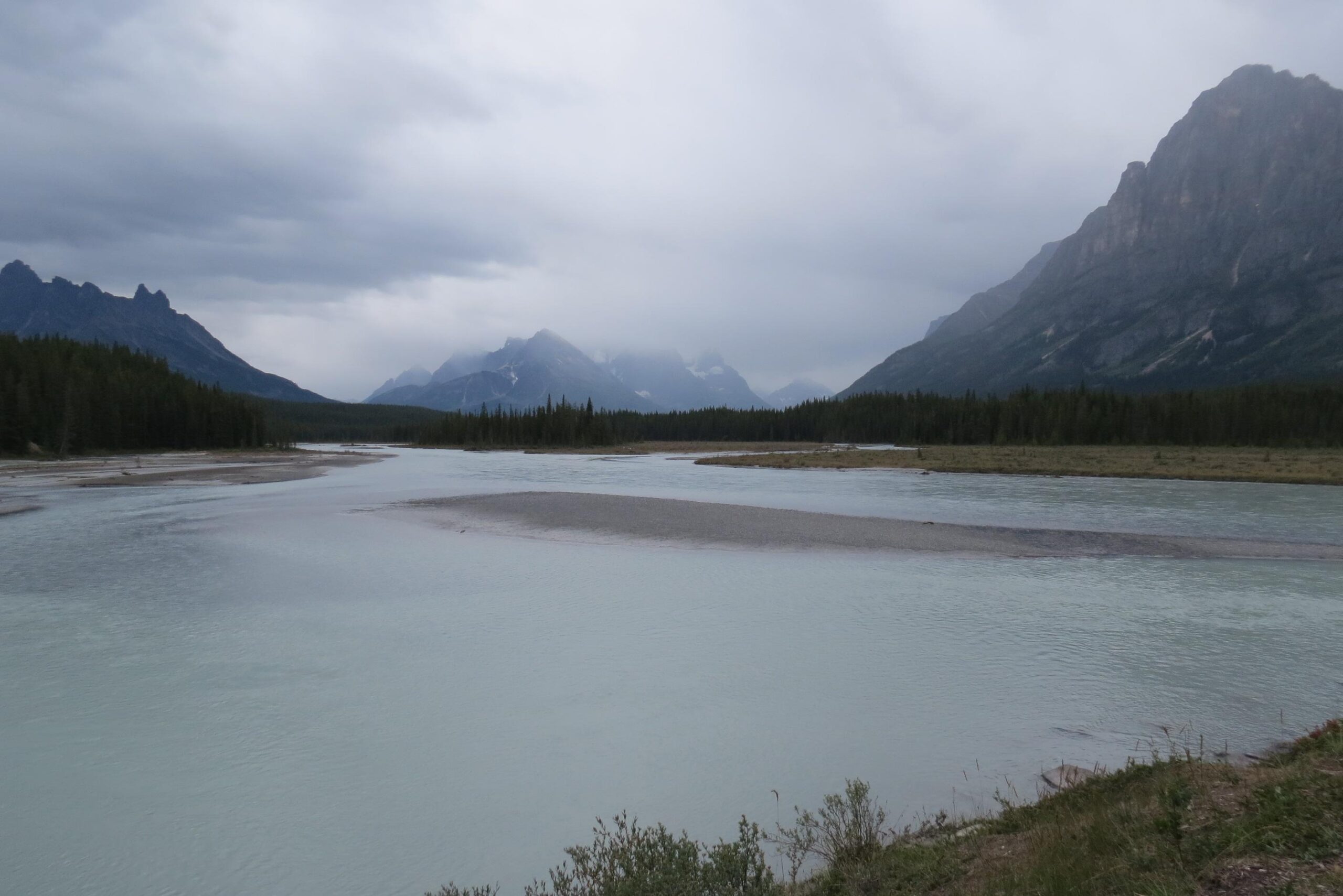 A serene landscape featuring a winding river surrounded by lush greenery and mountains under a cloudy sky. The water has a soft, turquoise hue and the distant mountains are partially shrouded in mist, creating a tranquil and atmospheric scene. Big Bend (Trail #126) mountain bike trail.