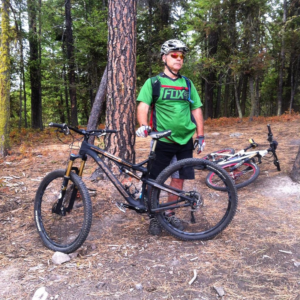 Intense 2014 Spider 29c: A mountain biker stands next to his bike in a forested area, wearing a green cycling jersey, helmet, and sunglasses. The background features tall trees and dry foliage, while another bike is positioned slightly behind him. The ground is covered with pine needles and rocks.