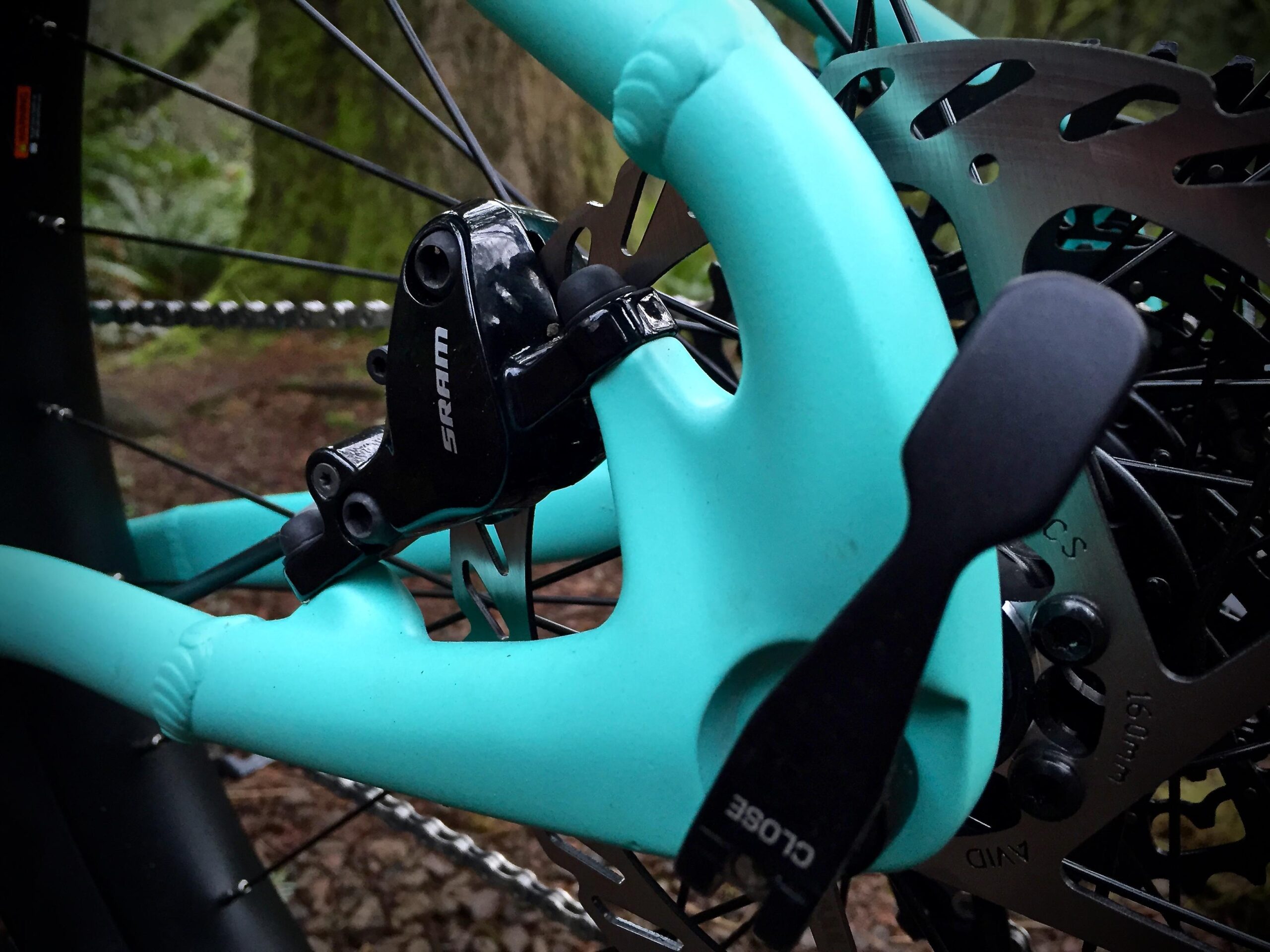 Norco Torrent 7: Close-up view of a bicycle's rear brake setup, featuring a turquoise frame, black SRAM brake caliper, and a disc brake rotor. The background shows a natural, wooded environment.