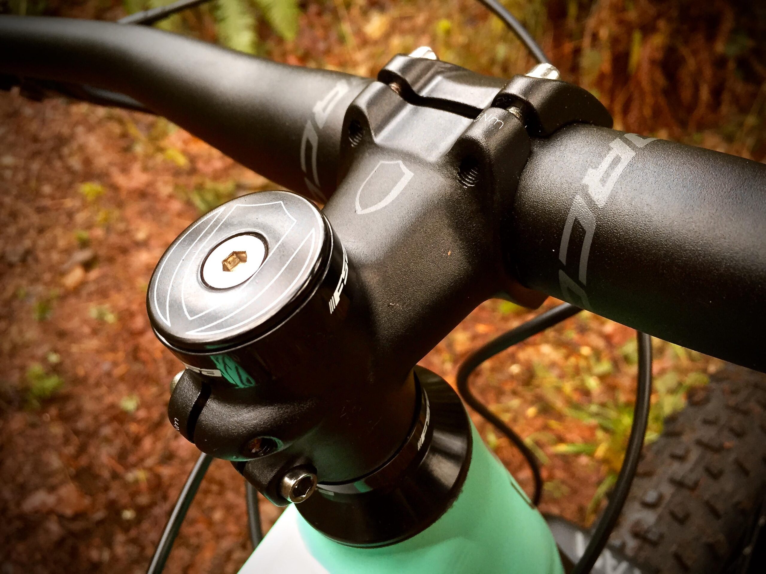 Norco Torrent 7: Close-up view of a bicycle handlebar and stem, featuring a black stem cap with a hexagonal socket, and a sleek black handlebar with subtle branding. The background shows earthy tones, indicating a natural outdoor setting.