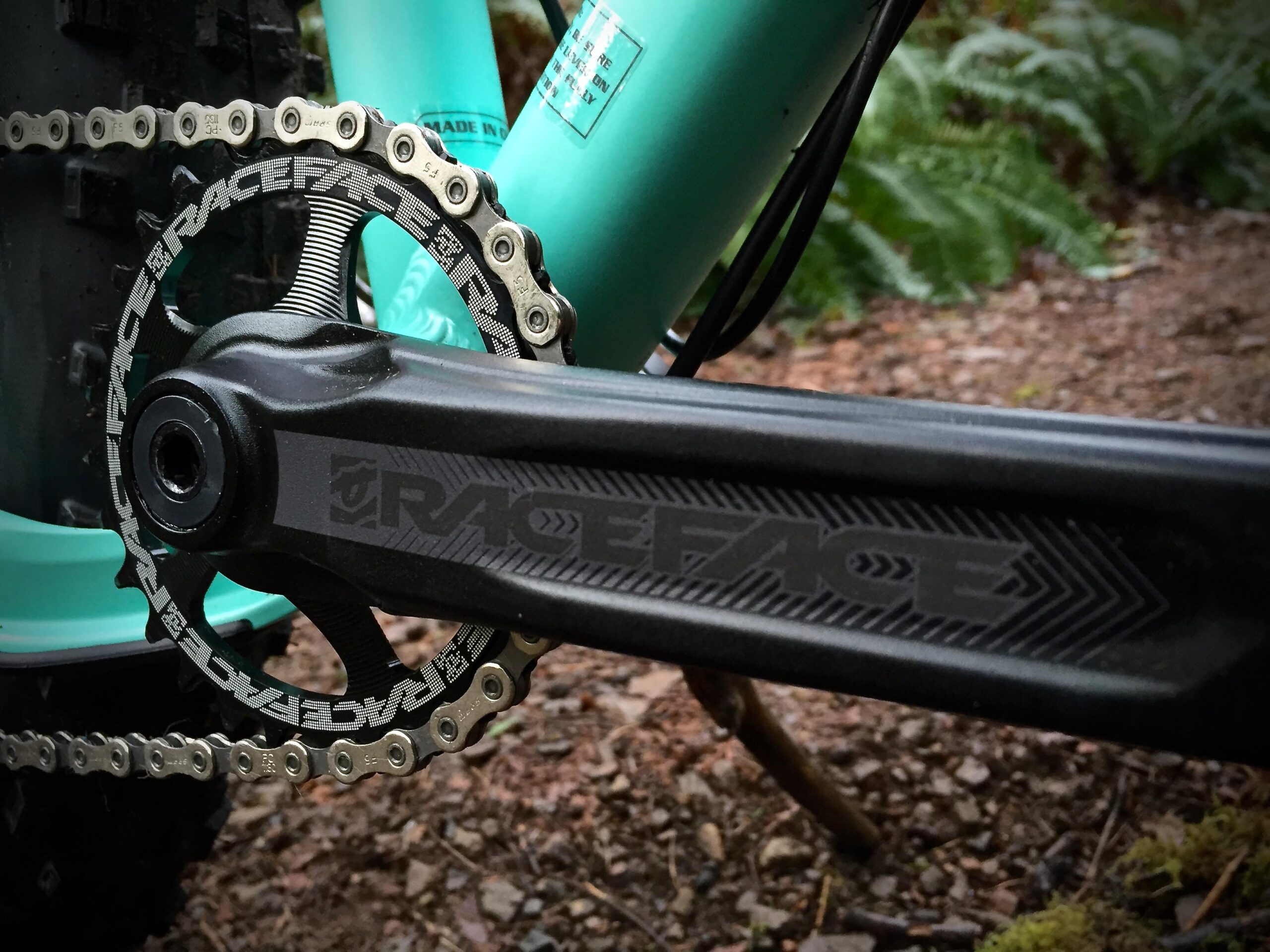 Norco Torrent 7: Close-up view of a bicycle's crankset featuring a black Race Face crank arm and chainring, with a silver chain in place. The background shows a turquoise bike frame and natural terrain including dirt and foliage.