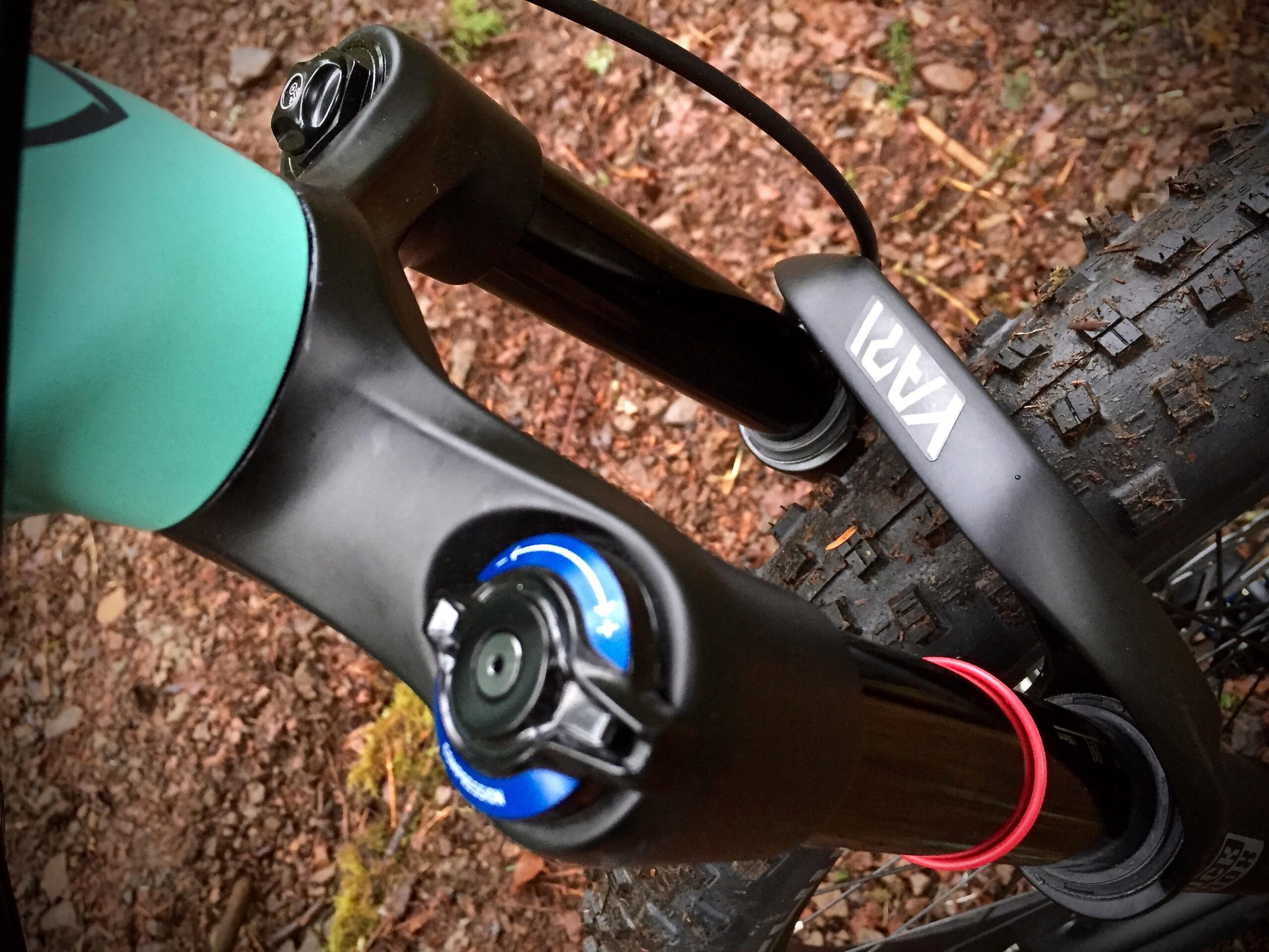 Norco Torrent 7: Close-up image of a bicycle's front suspension fork, featuring a blue dial for adjustment and a black and silver branding label. The fork is mounted above a tire with a textured tread, surrounded by gravel and natural elements.