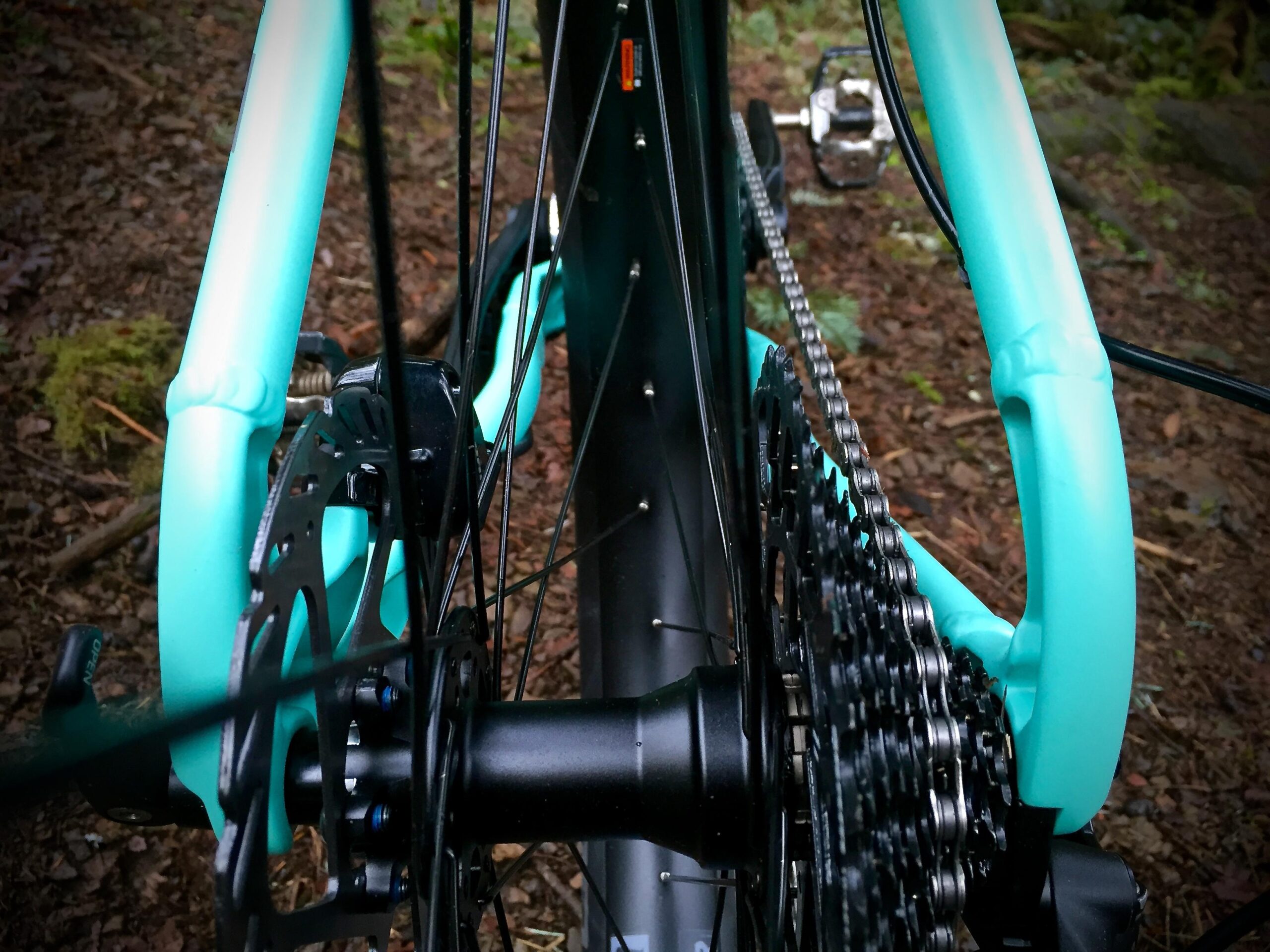 Norco Torrent 7: Close-up view of the rear wheel of a turquoise and black bicycle, highlighting the drivetrain, cassette, and disc brake against a natural, earthy background with leaves and moss.