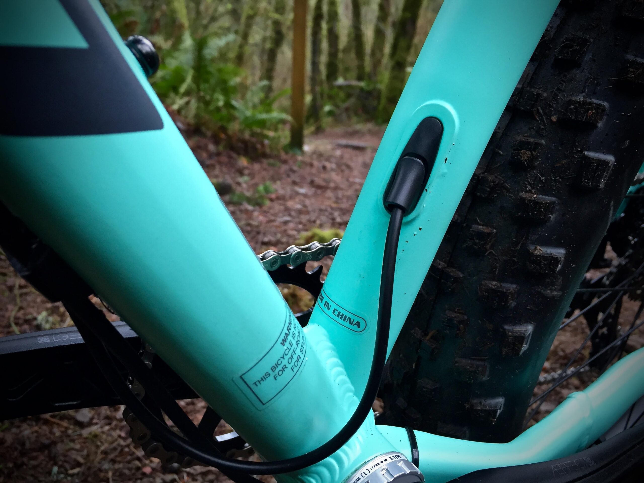 Norco Torrent 7: Close-up of a turquoise bicycle frame, highlighting the area where the cable is routed. The photograph captures the gear chain and a portion of a rugged tire, with a blurred forest background of trees and foliage.