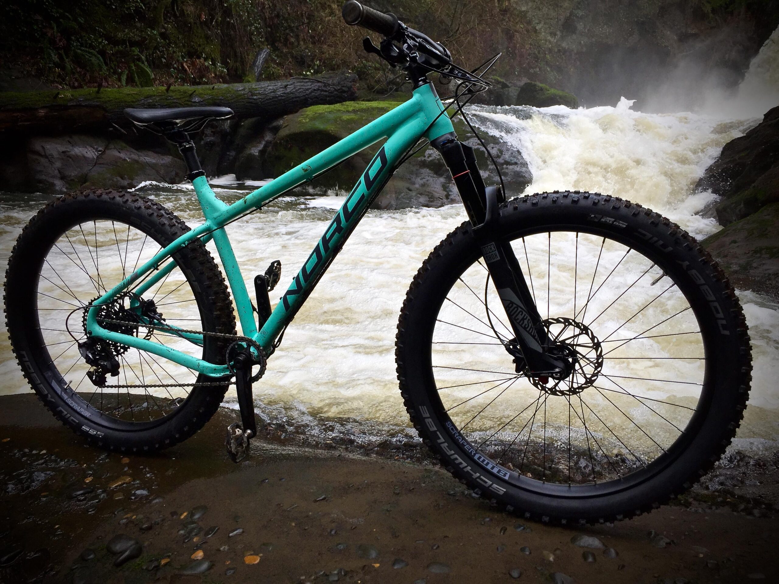 Norco Torrent 7: A turquoise mountain bike positioned on a rocky bank near a rushing waterfall. The bike features thick tires and a sturdy frame, with greenery in the background, emphasizing an outdoor adventure setting.