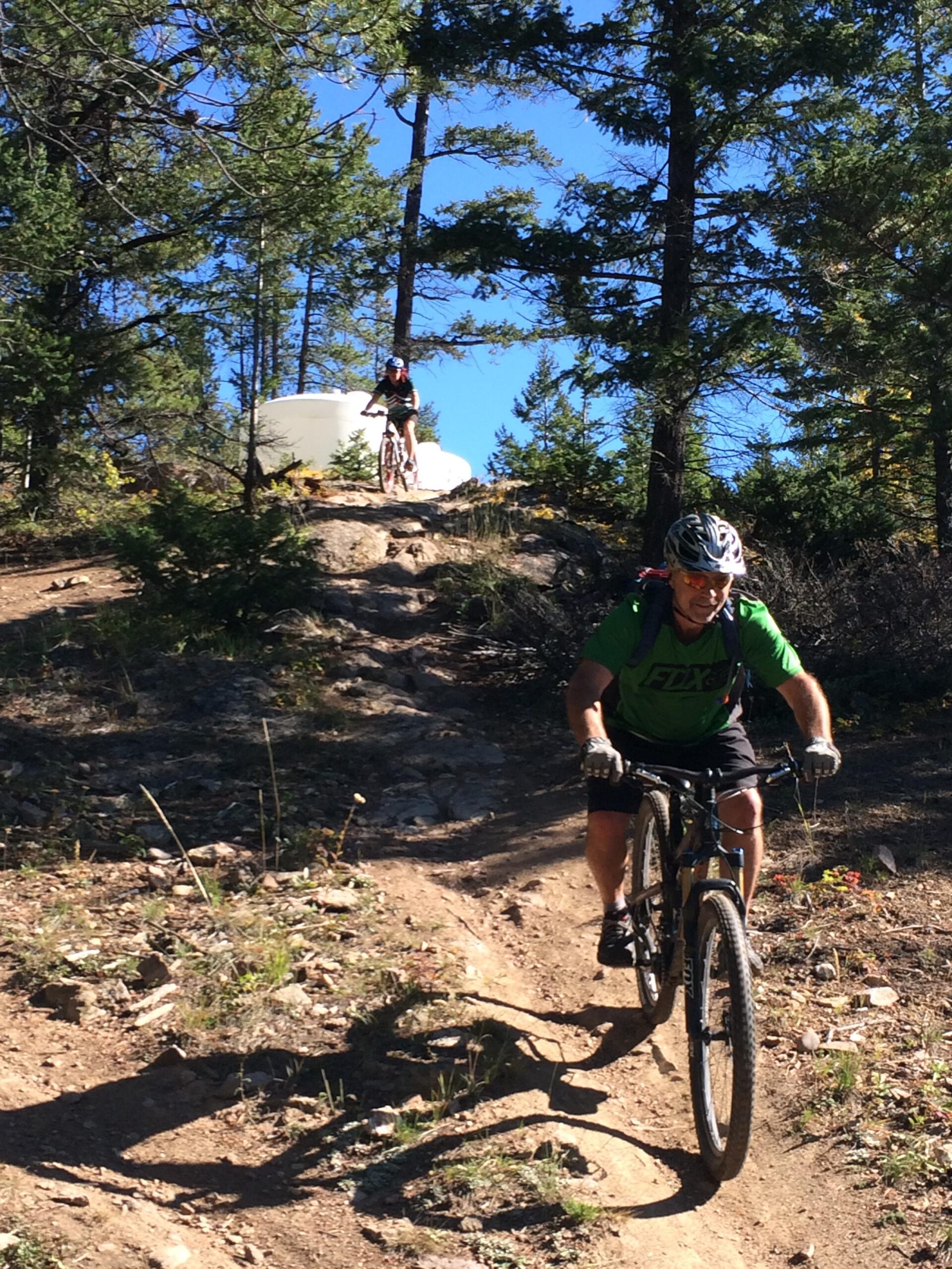 Two mountain bikers navigate a rocky trail in a forested area. One rider is ascending a steep incline, while another is positioned higher up on the trail. Surrounding are tall trees and a clear blue sky, indicating a sunny day. Watch Tower mountain bike trail.