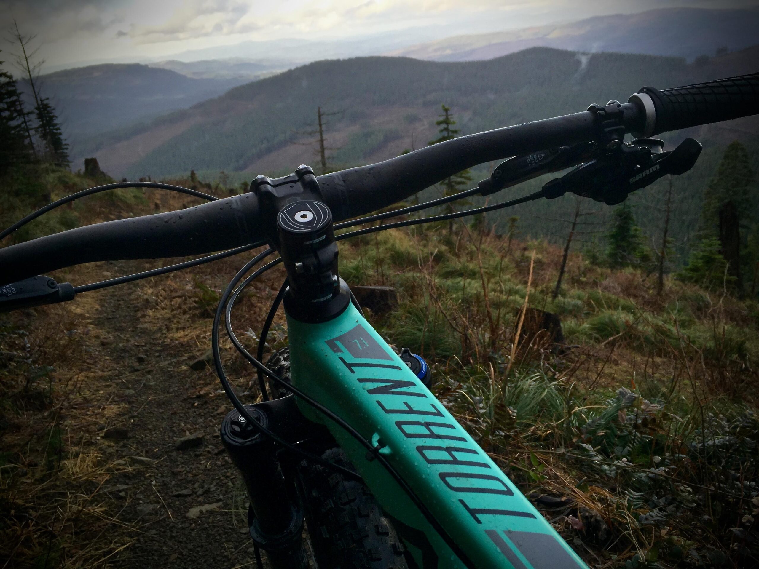 Norco Torrent 7: A close-up view of a mountain bike handlebars set against a scenic backdrop of rolling hills and dense forests. The image captures the bike's stem and brake lever, with rain droplets glistening on the surface. The pathway below is rocky and surrounded by greenery, suggesting an adventurous mountain biking trail.