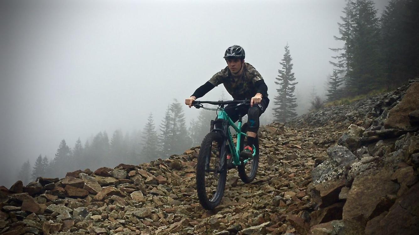 A mountain biker navigating a rocky trail through foggy terrain, surrounded by pine trees. The rider is wearing a helmet and camouflaged clothing, focused on maintaining balance on the challenging path. Cold Creek mountain bike trail.