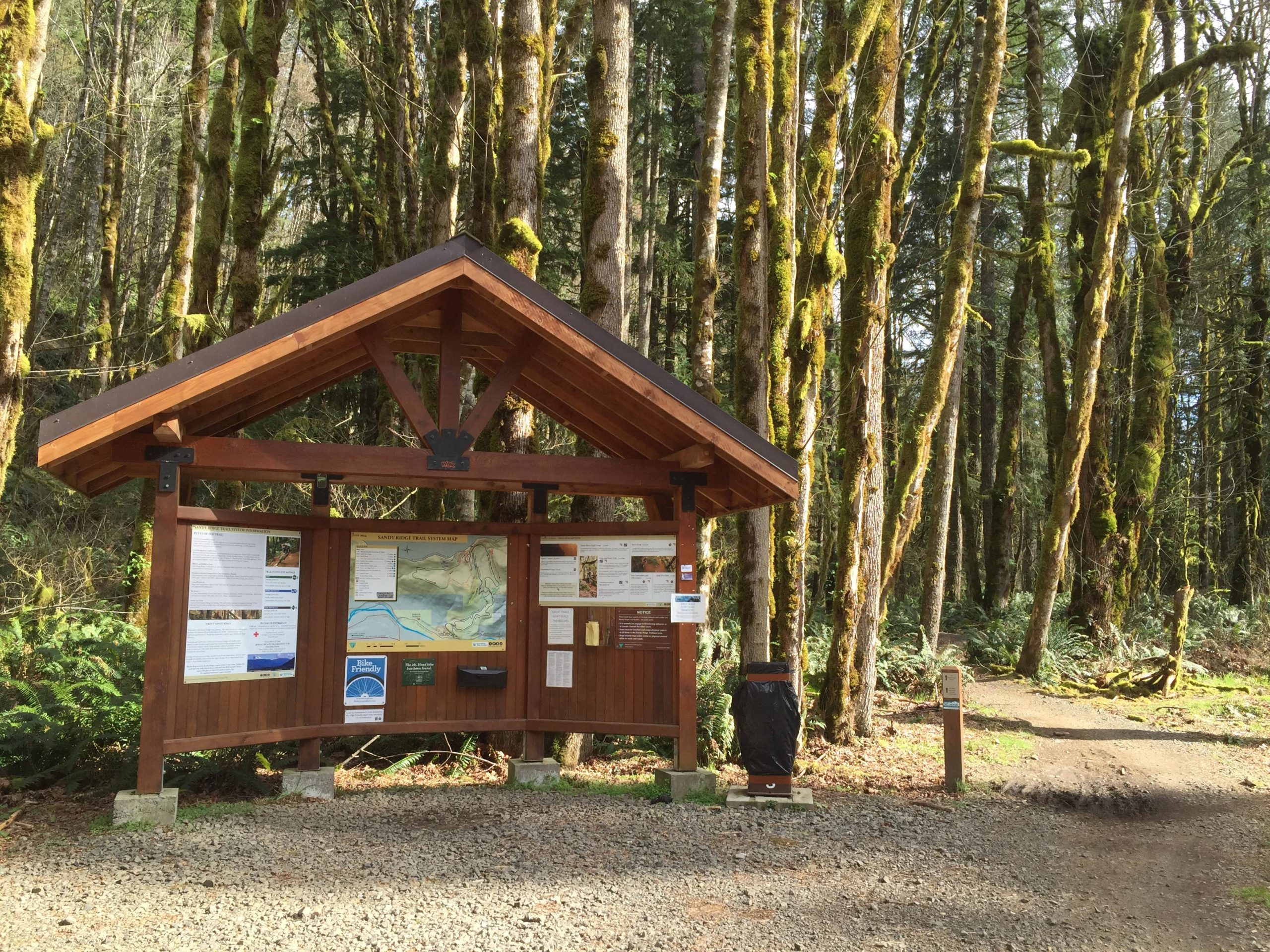 Wooden trailhead sign displaying maps and information in a lush, green forest. Tall trees with moss-covered trunks surround the sign, and a gravel path leads into the woods. Sandy Ridge mountain bike trail.