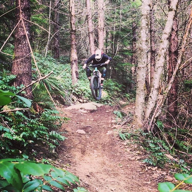 A mountain biker performing a jump on a dirt trail surrounded by trees and greenery. The rider is elevated off the ground, captured in mid-air with focused determination. The trail features rocky terrain and lush foliage. Sandy Ridge mountain bike trail.