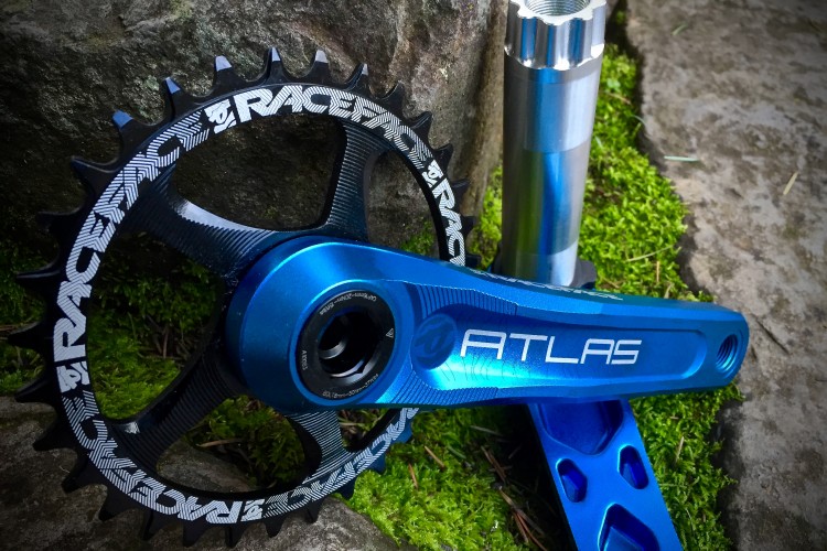 A close-up view of a blue bicycle crankset featuring a chainring with visible branding. The crank arm is positioned on a natural surface with stones and moss in the background, showcasing the details of the components.