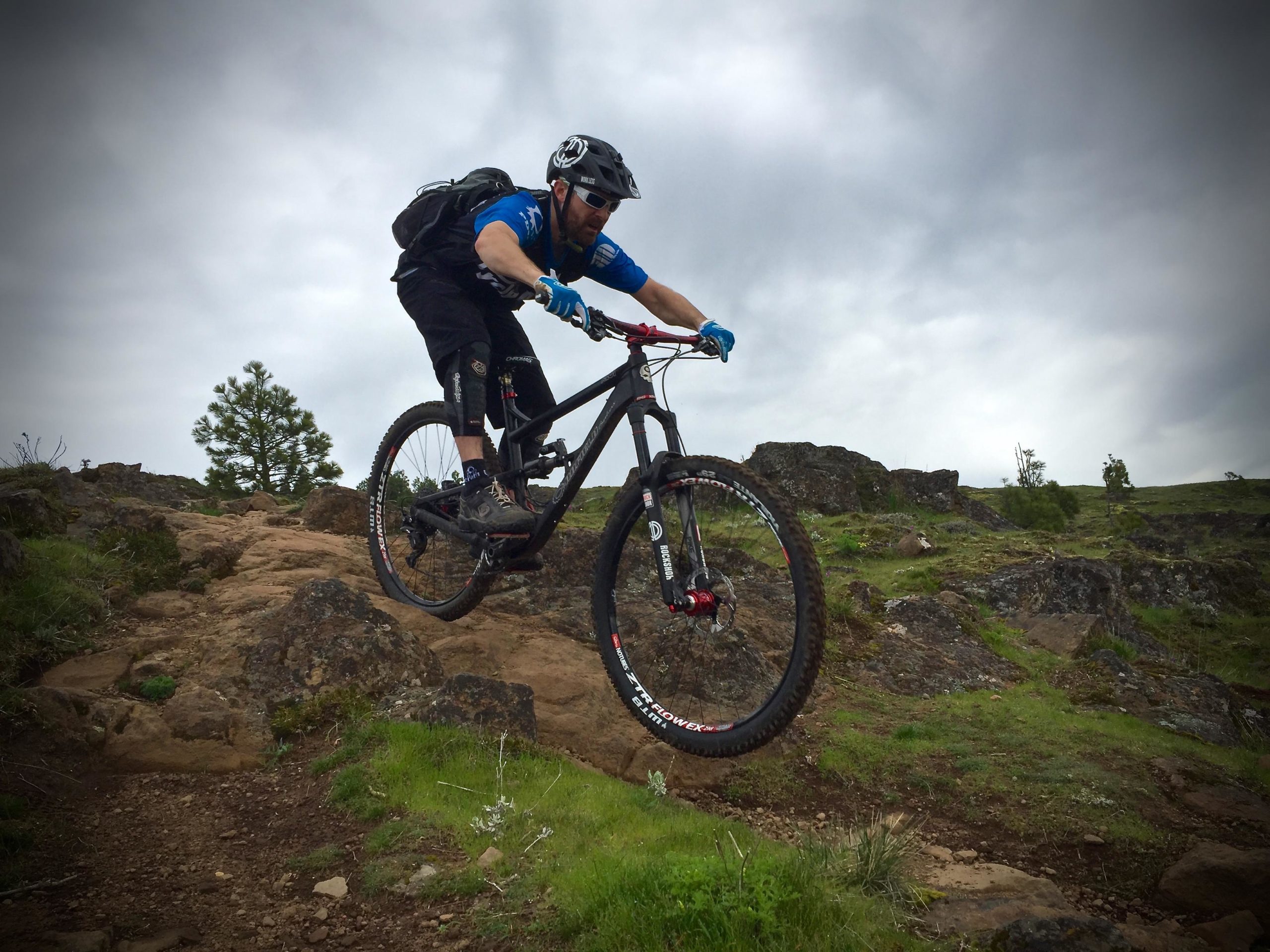 A mountain biker in a blue shirt and helmet is skillfully navigating rocky terrain, with one wheel off the ground as he jumps over a boulder. The landscape features green grass and scattered rocks under a cloudy sky. Syncline mountain bike trail.