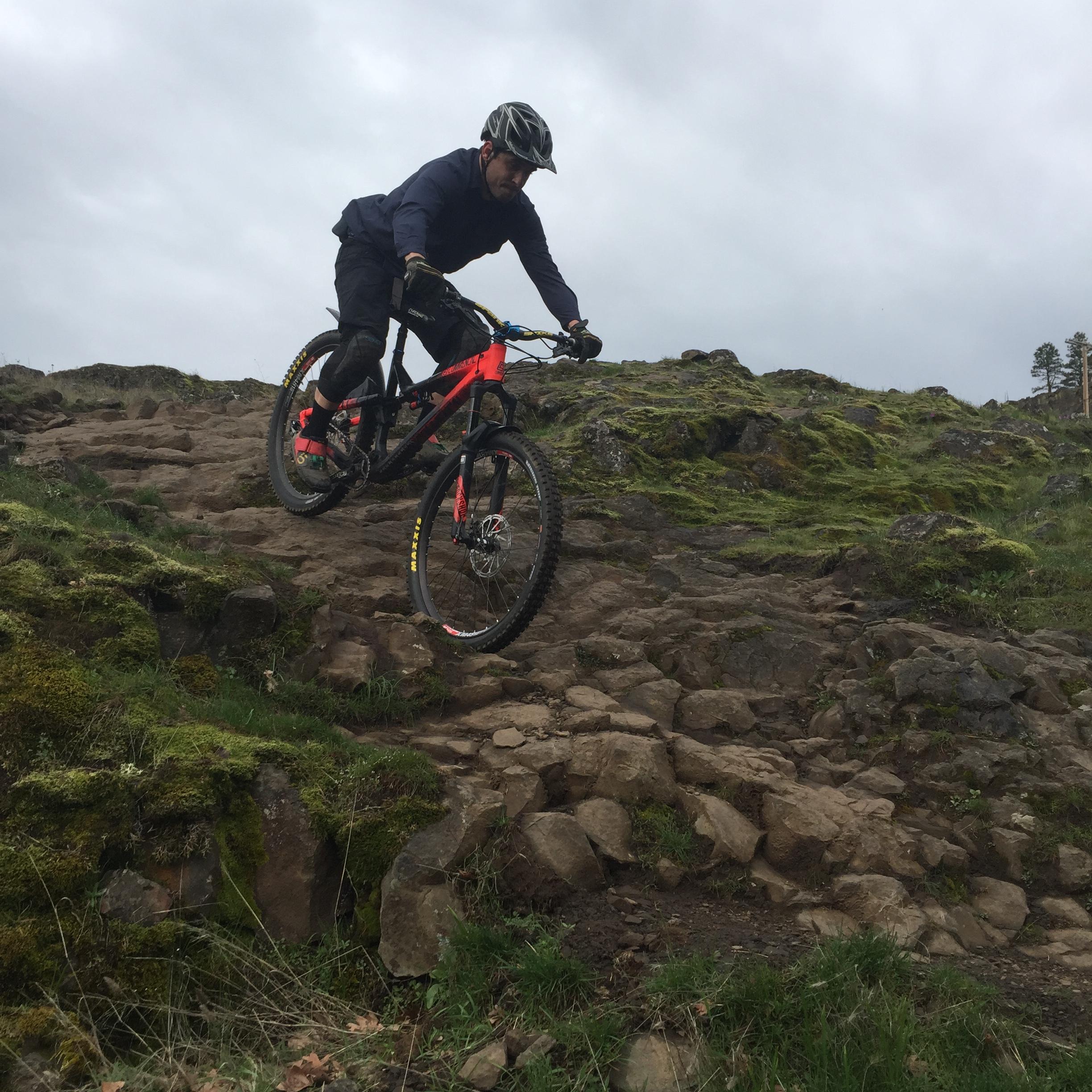 Commencal Meta AM V4: A mountain biker in a helmet and protective gear descends a rocky trail, skillfully navigating the terrain on a red and black mountain bike. Surrounding the scene are patches of green grass and moss, under a cloudy sky.