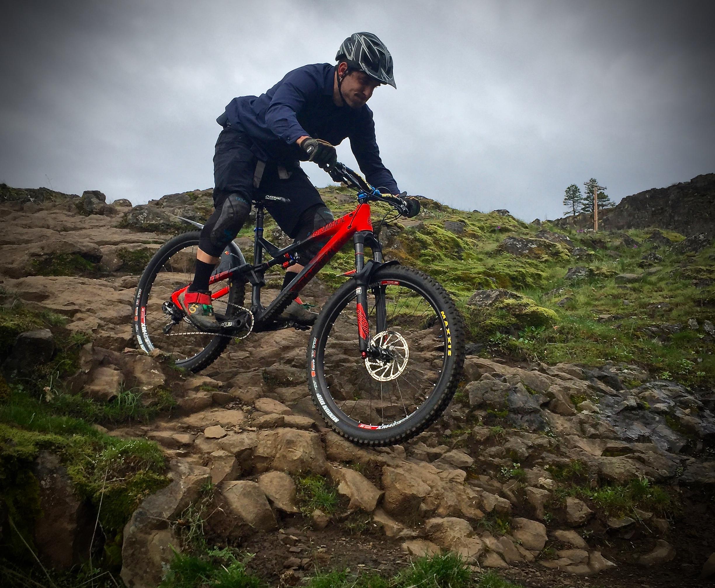 A mountain biker descending a rocky trail, wearing a helmet and protective gear, on a red mountain bike. The background features rugged terrain and sparse greenery under a cloudy sky. Syncline mountain bike trail.