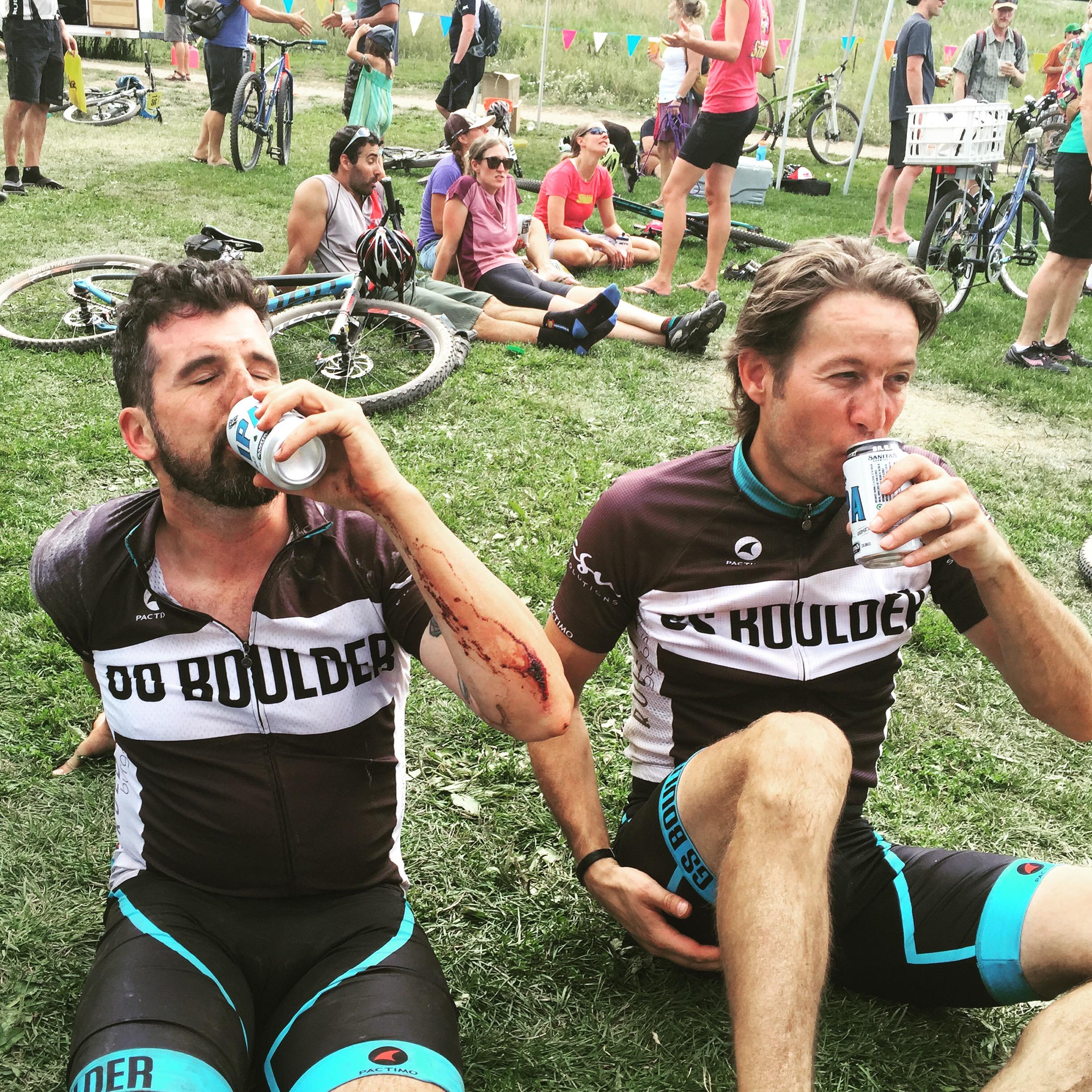 Two cyclists sitting on the grass, both wearing matching black and white jerseys that read "DO BOULDER." One cyclist has a visible injury on his arm and is holding a can of beer while leaning back. The other cyclist is positioned next to him, also drinking a can. In the background, other individuals and bicycles can be seen, indicating an outdoor event or gathering.