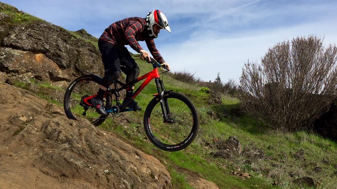 A mountain biker in a helmet and protective gear jumps off a rocky outcrop, showcasing skill and excitement. The bike is red with black accents, and the background features green grass and sparse vegetation under a blue sky. Syncline mountain bike trail.