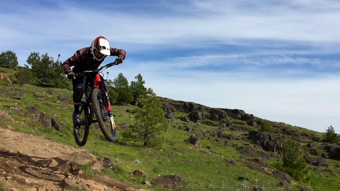 A mountain biker in a helmet and protective gear performs a jump on a dirt trail surrounded by green grass and rocky terrain under a blue sky. Syncline mountain bike trail.