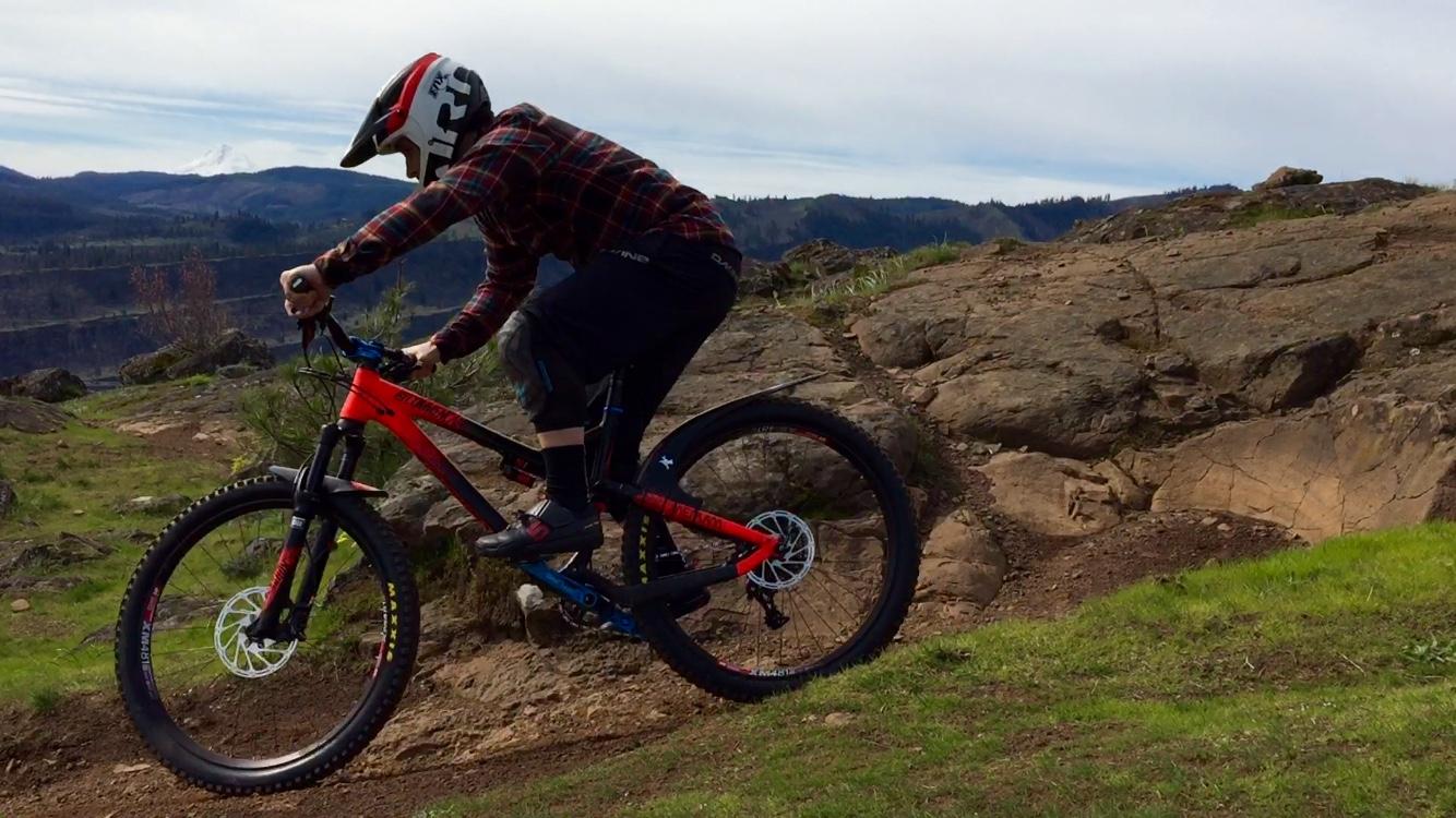 A mountain biker in a helmet and a plaid shirt performs a jump on a rocky trail surrounded by green grass and distant mountains under a cloudy sky. Syncline mountain bike trail.