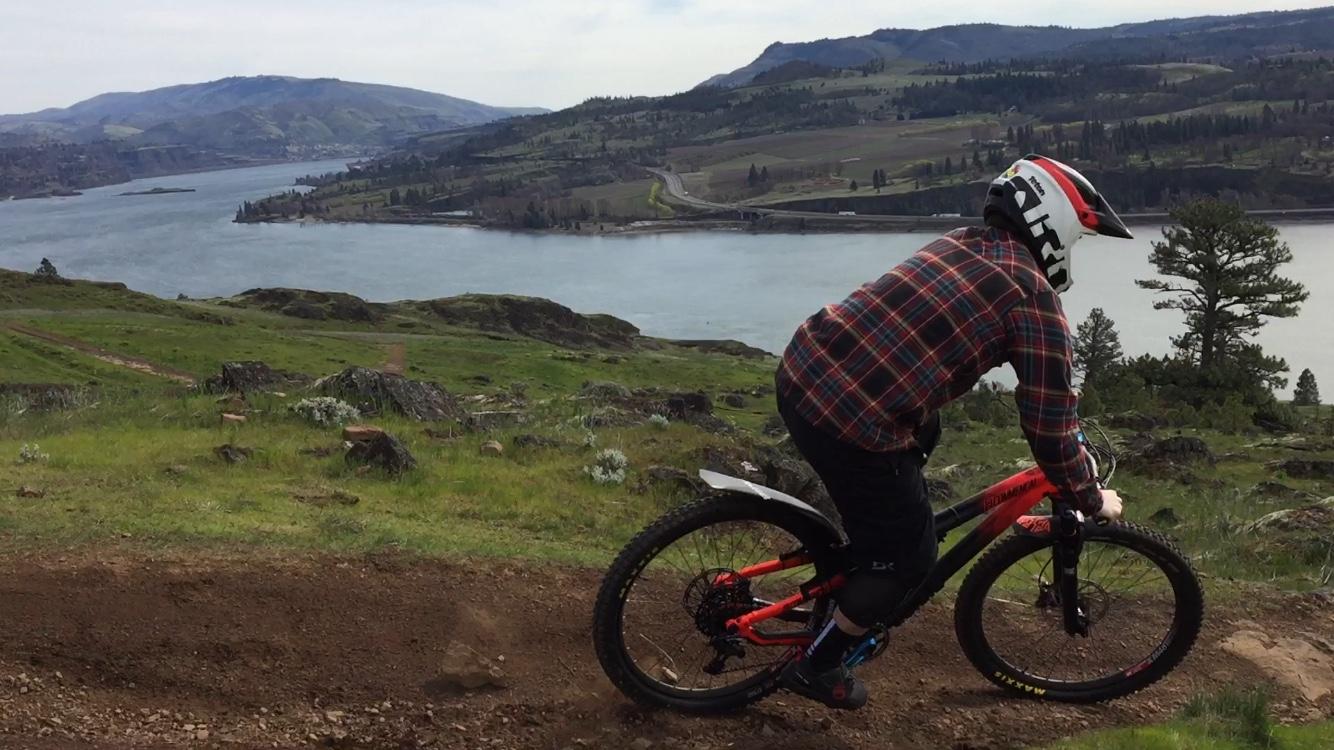 A mountain biker wearing a helmet and plaid shirt navigates a dirt trail on a downhill slope, with a scenic view of a river and lush green hills in the background. Syncline mountain bike trail.