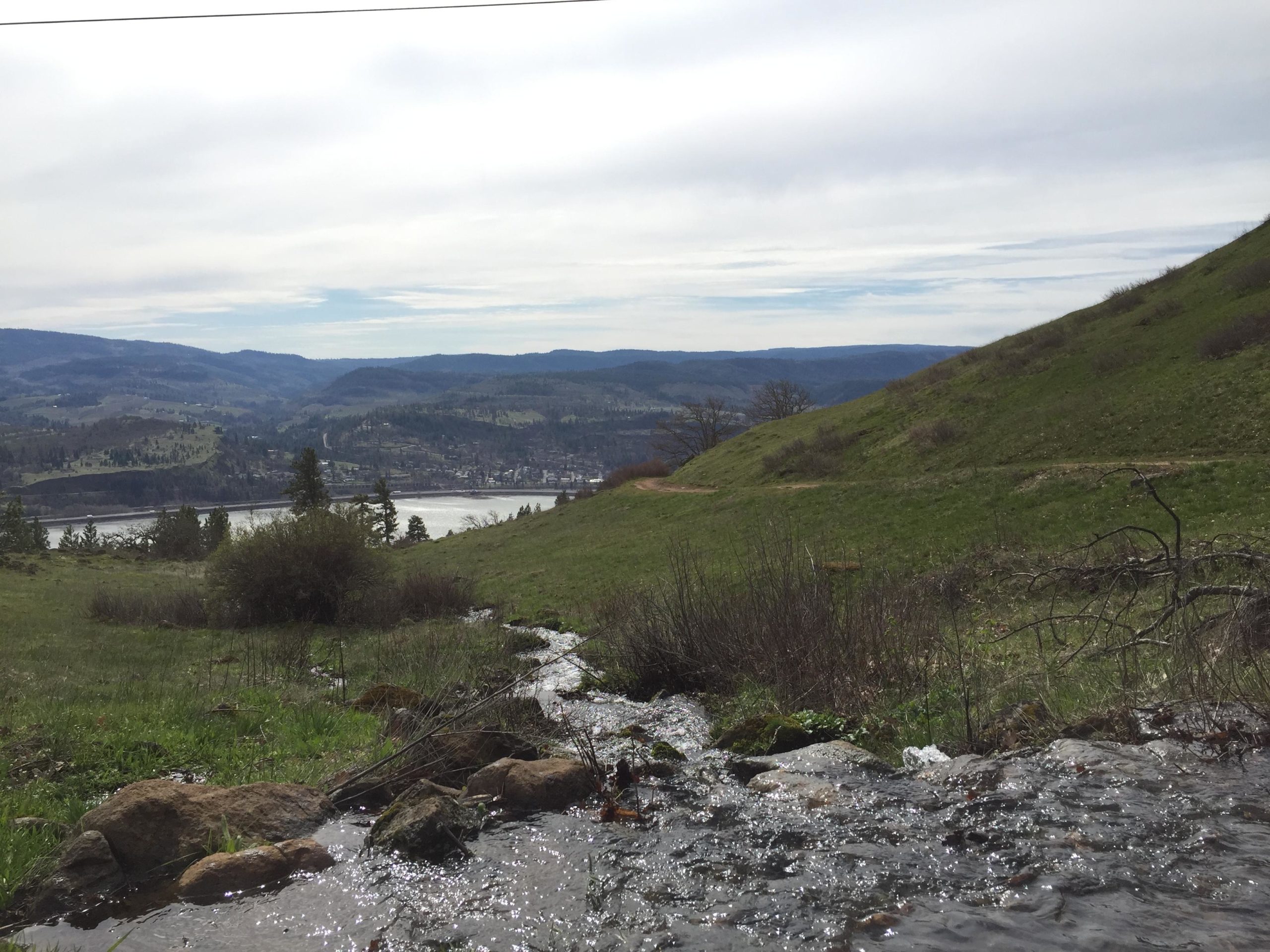 A scenic landscape featuring a gently flowing stream in the foreground, surrounded by green grass and shrubs. Rolling hills and mountainous terrain are visible in the background, along with a town and a body of water reflecting the sky. The sky is overcast, creating a serene atmosphere. Syncline mountain bike trail.