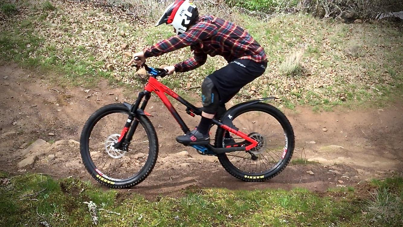 A mountain biker in a red and black plaid shirt and protective gear is riding a red mountain bike along a dirt trail in a forested area. The bike is mid-jump, showcasing its suspension as the rider leans forward, focused on maintaining balance. Surrounding the trail is lush greenery and scattered leaves, indicating a natural outdoor setting. Syncline mountain bike trail.