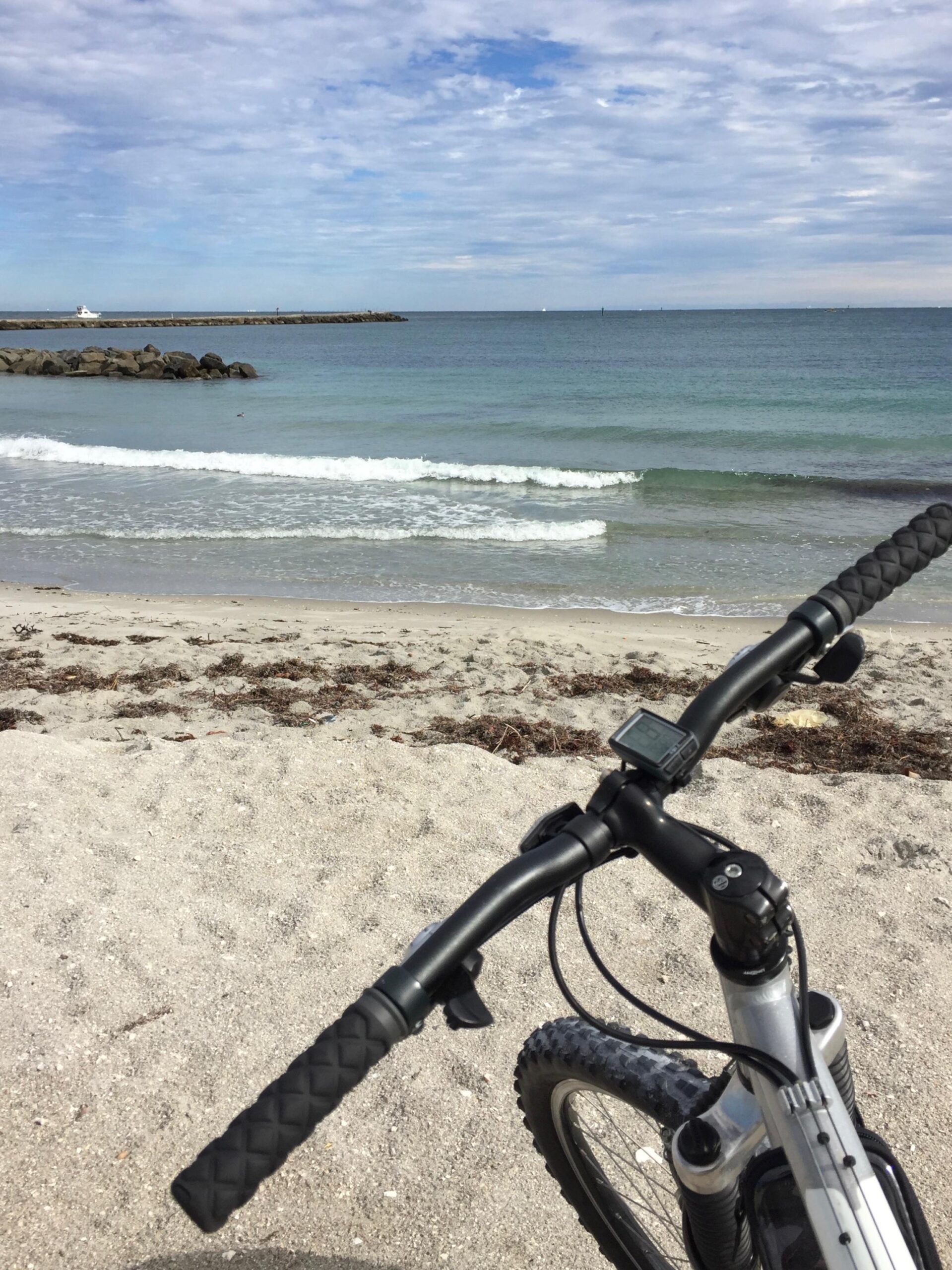 Gary Fisher Marlin: A close-up view of a bicycle's handlebars positioned on a sandy beach, with gentle waves lapping at the shore in the background. The scene features a partly cloudy sky and a rocky jetty extending into the water in the distance.