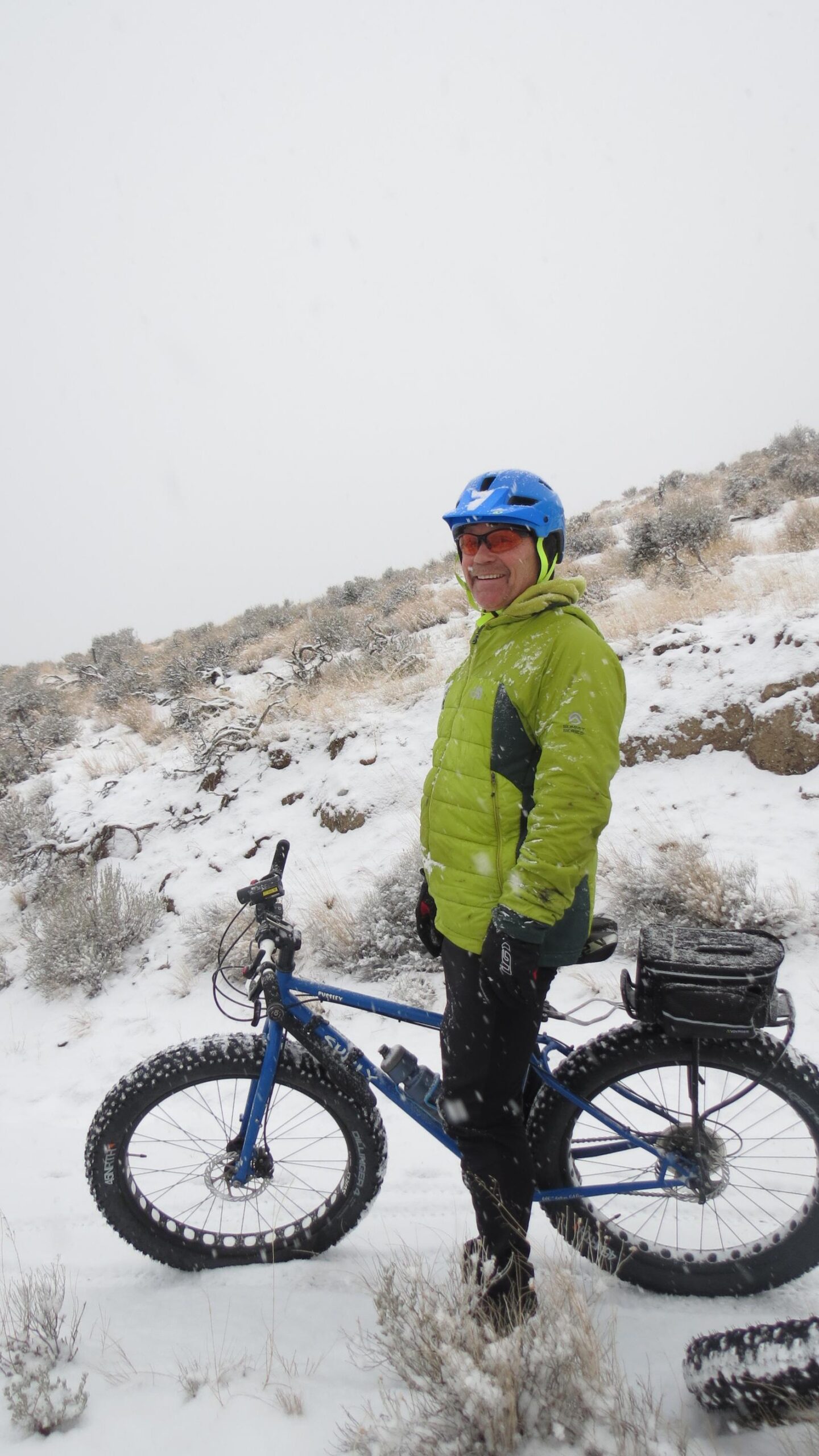 Scott Big Ed: A person wearing a bright green jacket and a blue helmet stands next to a blue fat bike on a snow-covered trail. Light snowflakes are falling, and the surrounding landscape features sparse vegetation and rocky terrain. The individual is smiling and appears to be enjoying the winter weather.