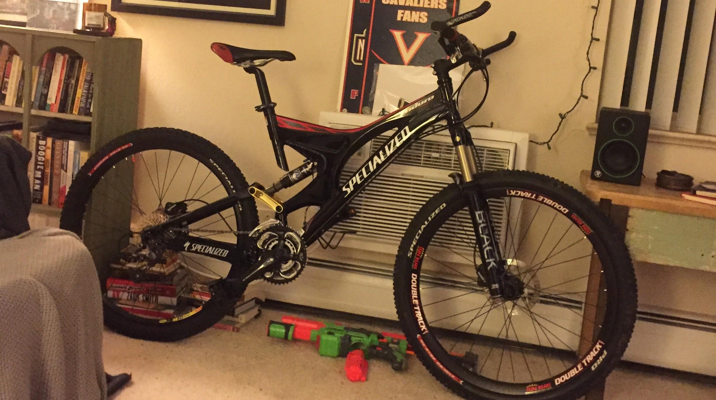 Specialized Enduro: A black Specialized mountain bike with red and gold accents is positioned in a living room setting. The bike is leaning against a wall near an air conditioning unit, with partially visible books stacked on the floor beneath it. In front of the bike, there are some colorful toy guns scattered on the carpet. An open space on the side features a small speaker and a shelf with books.