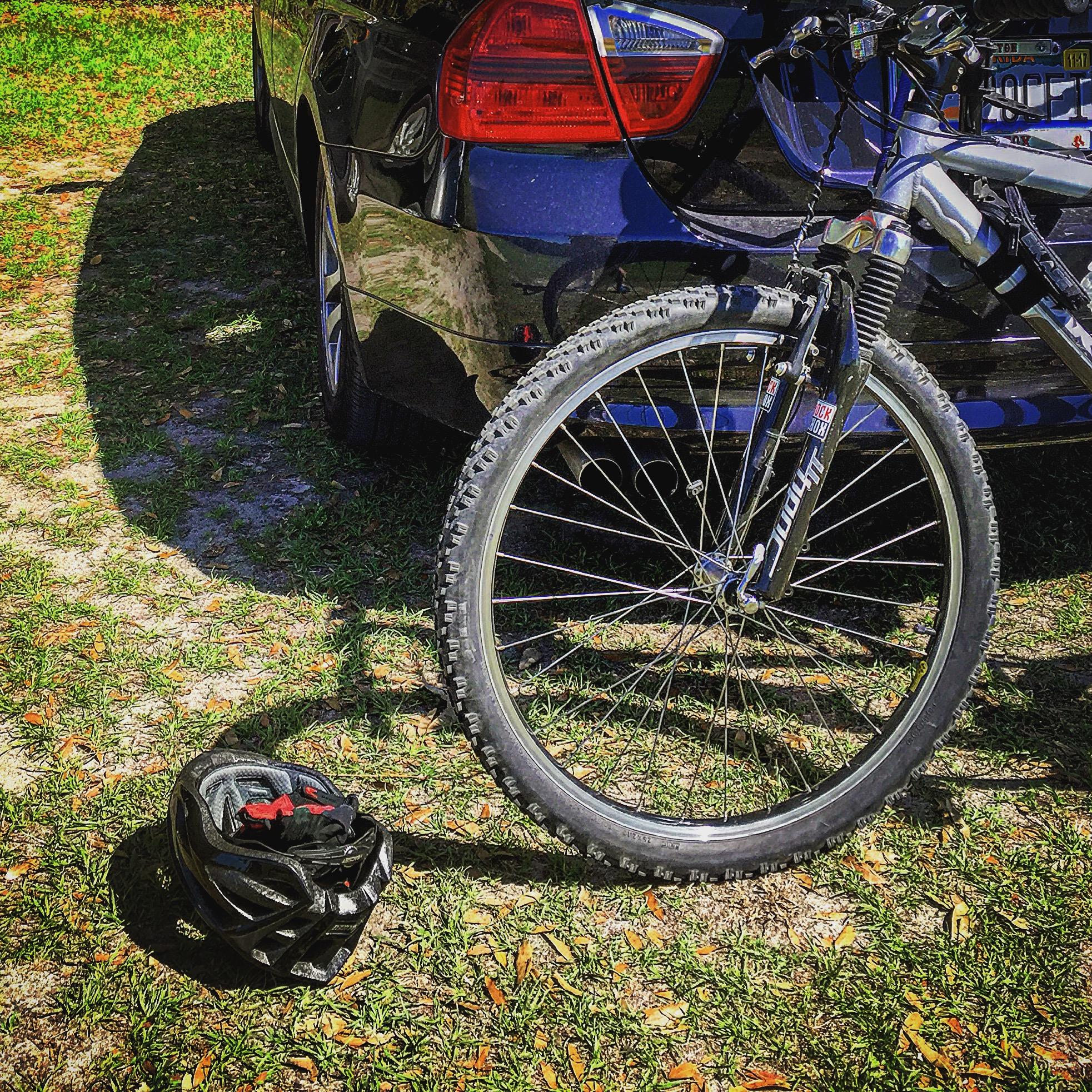 A mountain bike leaning against a black car, with a bicycle helmet lying on the ground beside it. The scene is set on a grassy area, showcasing the bike's front wheel and tire details. San Felasco Hammock Preserve mountain bike trail.