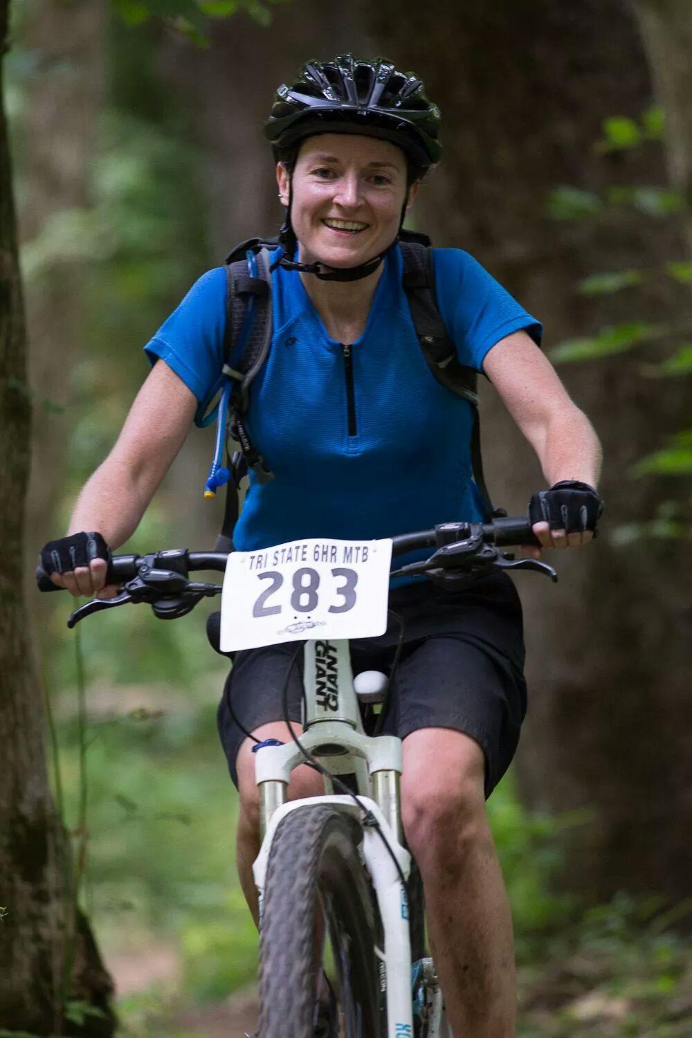 A smiling female mountain biker wearing a blue shirt and a helmet is riding a bike on a wooded trail. The bike has a number plate displaying "283" attached to the front. The background features lush green trees, indicating an outdoor setting. England Idlewild Mountain Biking Park mountain bike trail.