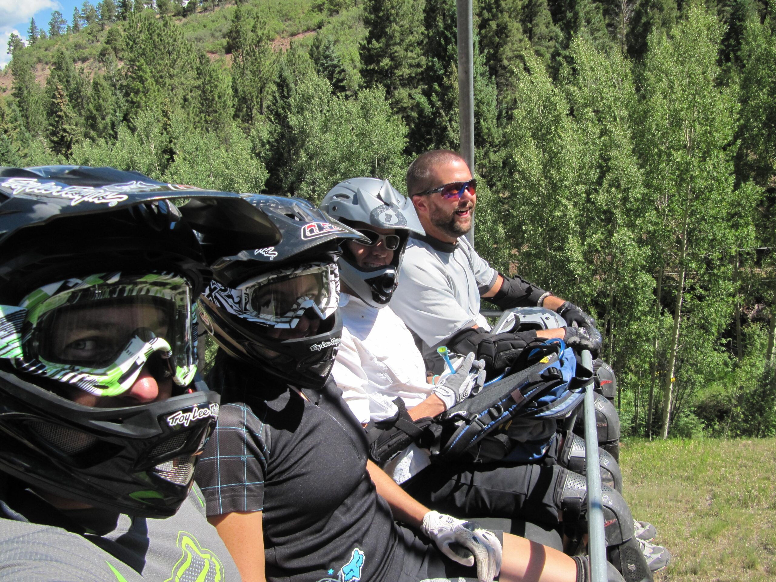 A group of four individuals wearing protective gear and helmets are seated on a lift, preparing for a mountain biking adventure. They are surrounded by green trees and a sunny sky, with visible smiles reflecting excitement. Angel Fire Bike Park mountain bike trail.