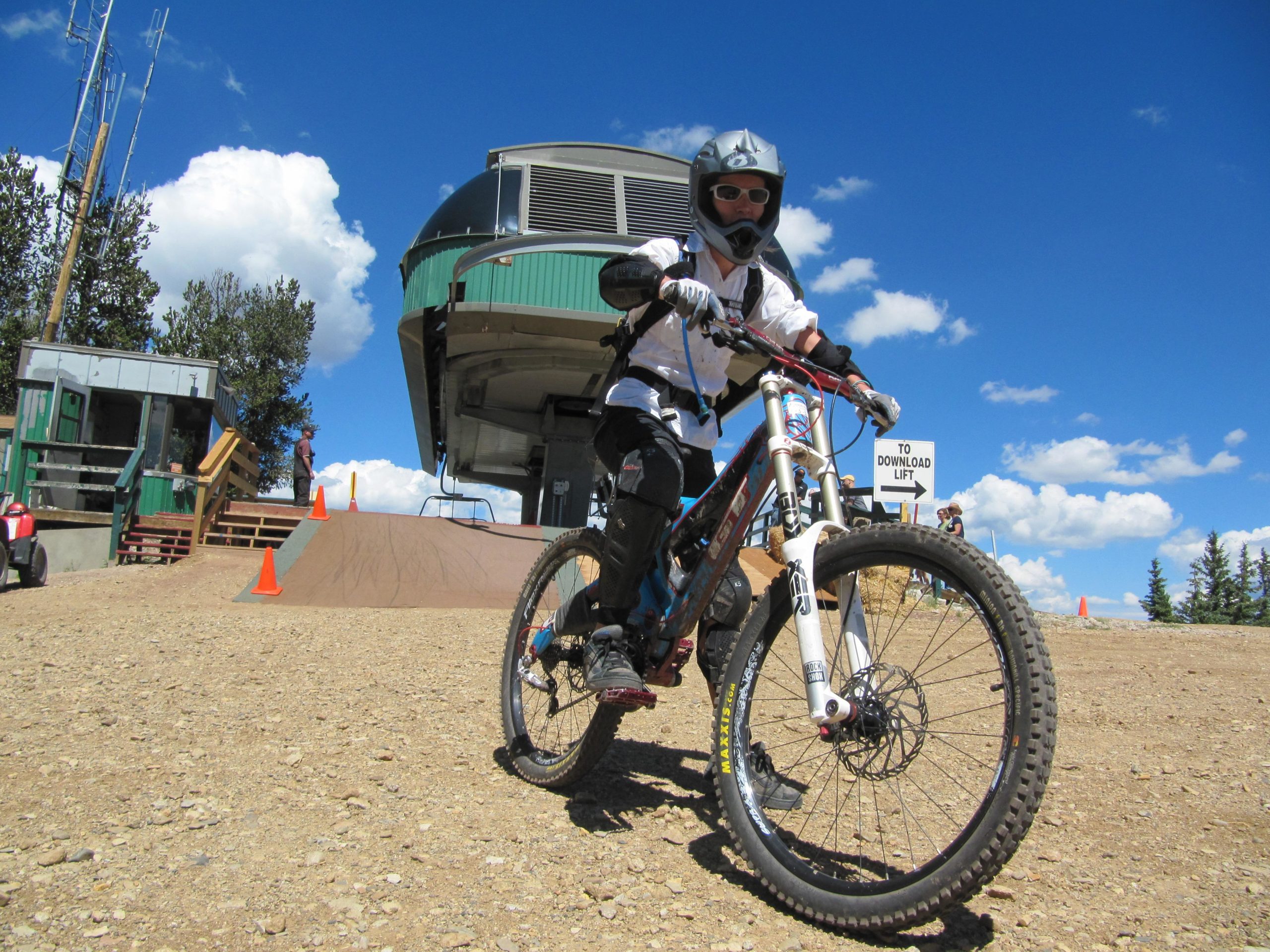 A cyclist wearing a helmet and protective gear is preparing to ride down a dirt ramp at a mountain biking park. In the background, there is a ski lift with a green and silver gondola, surrounded by trees and blue skies with fluffy white clouds. Safety cones are positioned along the ramp, and a sign indicates the direction to the download lift. Angel Fire Bike Park mountain bike trail.