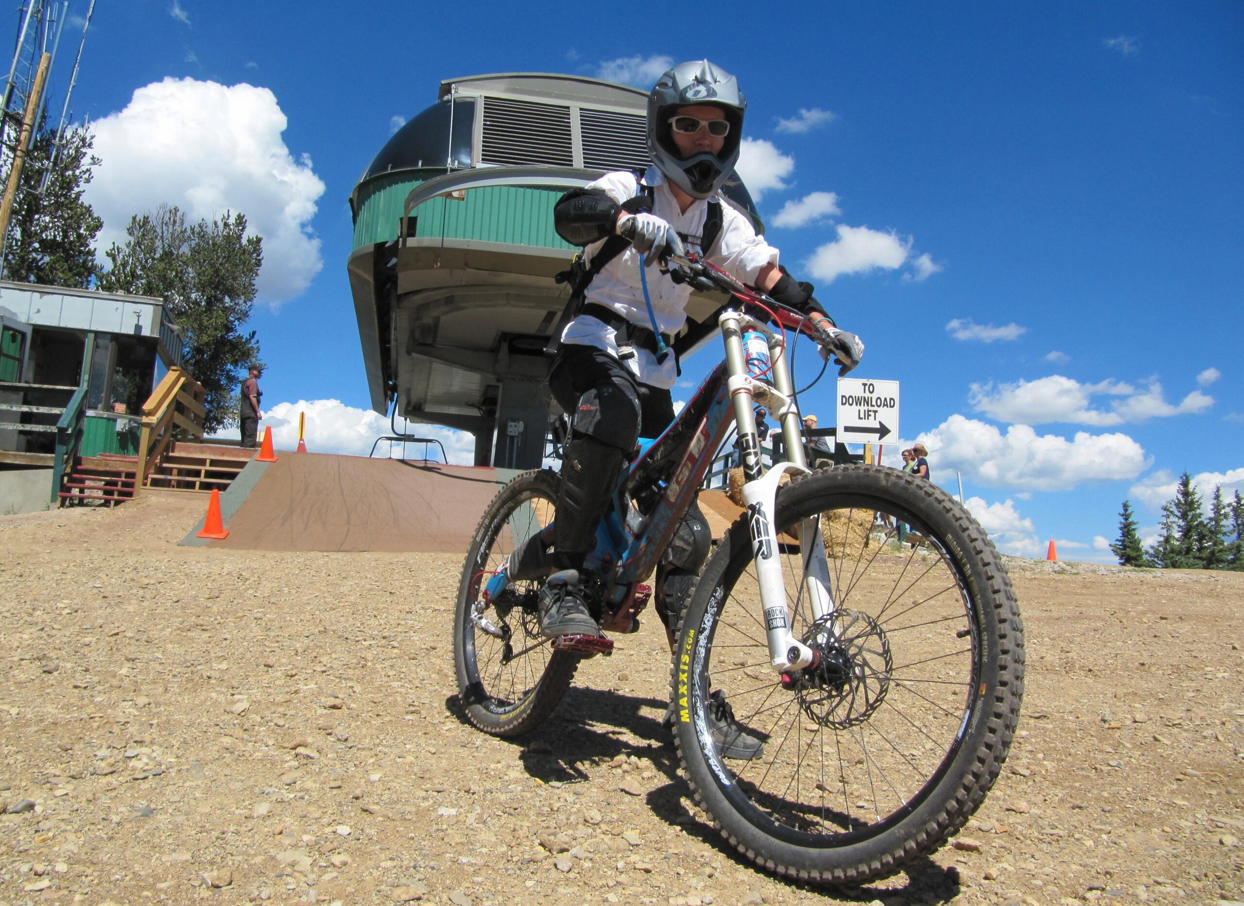 A young mountain biker in protective gear, including a helmet and gloves, is preparing to ride down a dirt ramp at a ski resort. In the background, a chairlift is visible alongside a sign indicating the direction to the download lift. The sky is bright blue with scattered clouds. Angel Fire Bike Park mountain bike trail.