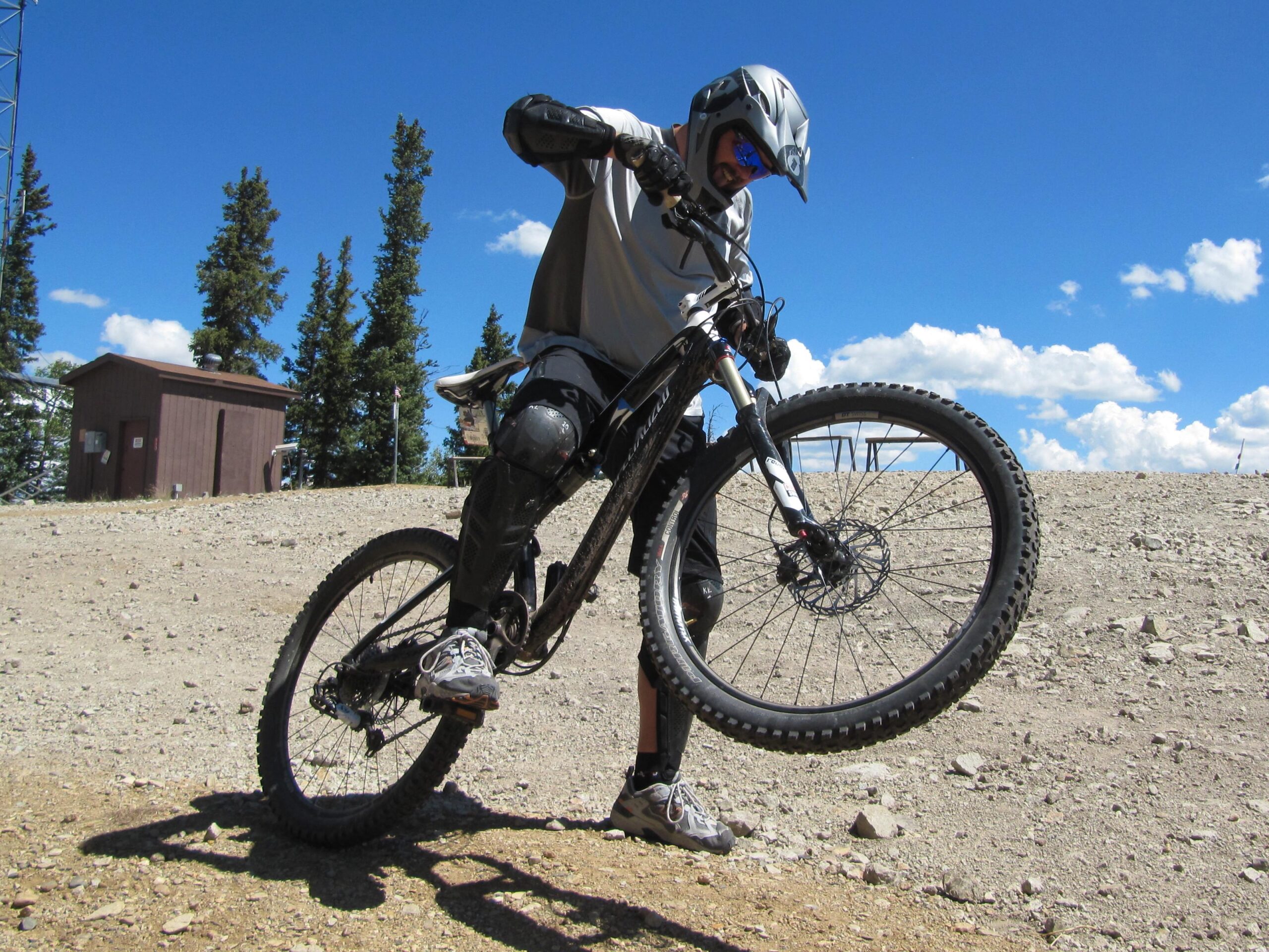 A mountain biker performing a stunt by lifting the front wheel of their bike while on a dirt trail. The rider is wearing a helmet and protective gear, and there are trees and a blue sky with clouds in the background. Angel Fire Bike Park mountain bike trail.