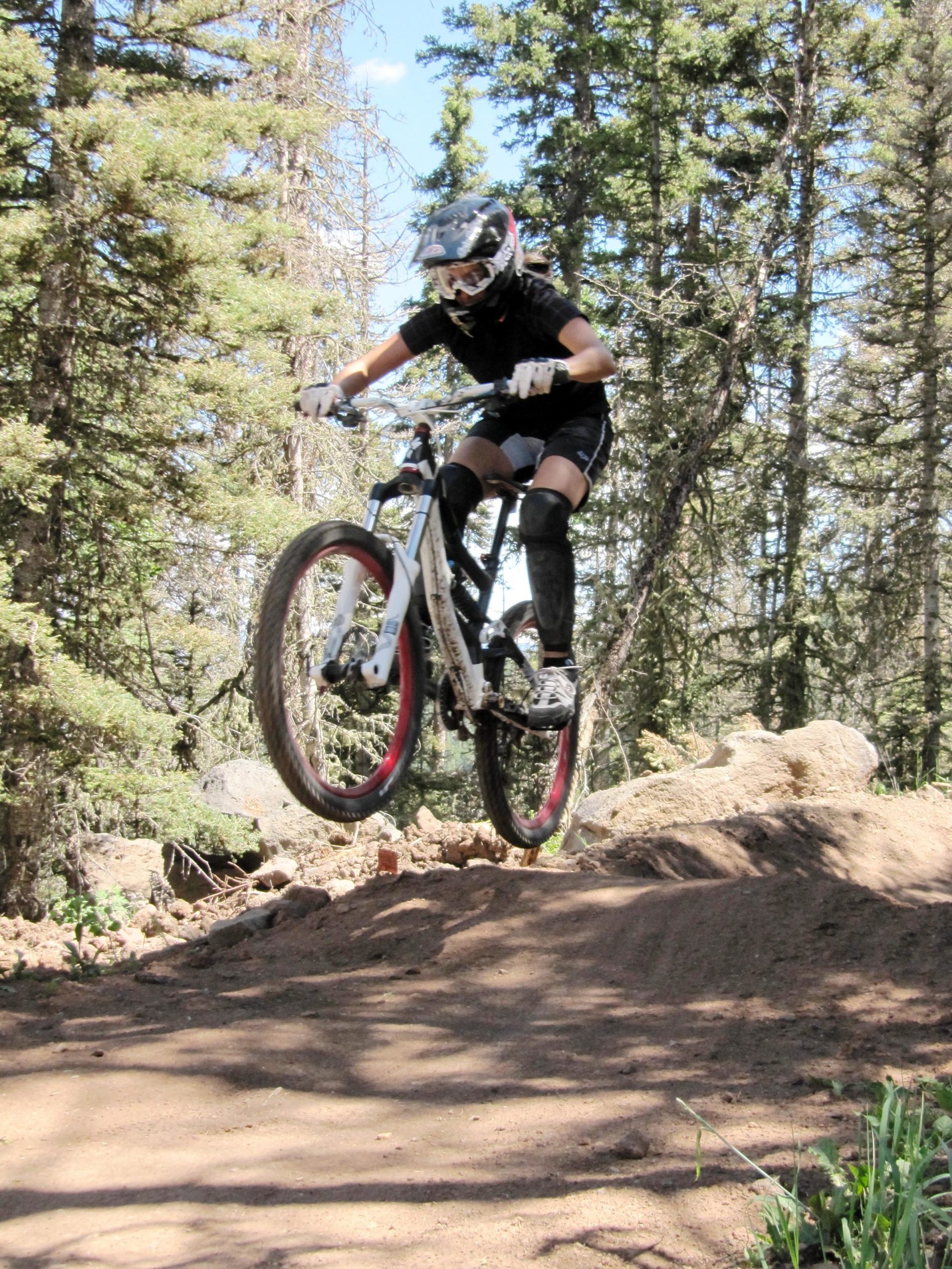 A person in protective cycling gear is mid-air while riding a mountain bike on a dirt trail surrounded by trees. The cyclist has a focused expression and is landing from a jump, showcasing an active outdoor scene. Angel Fire Bike Park mountain bike trail.