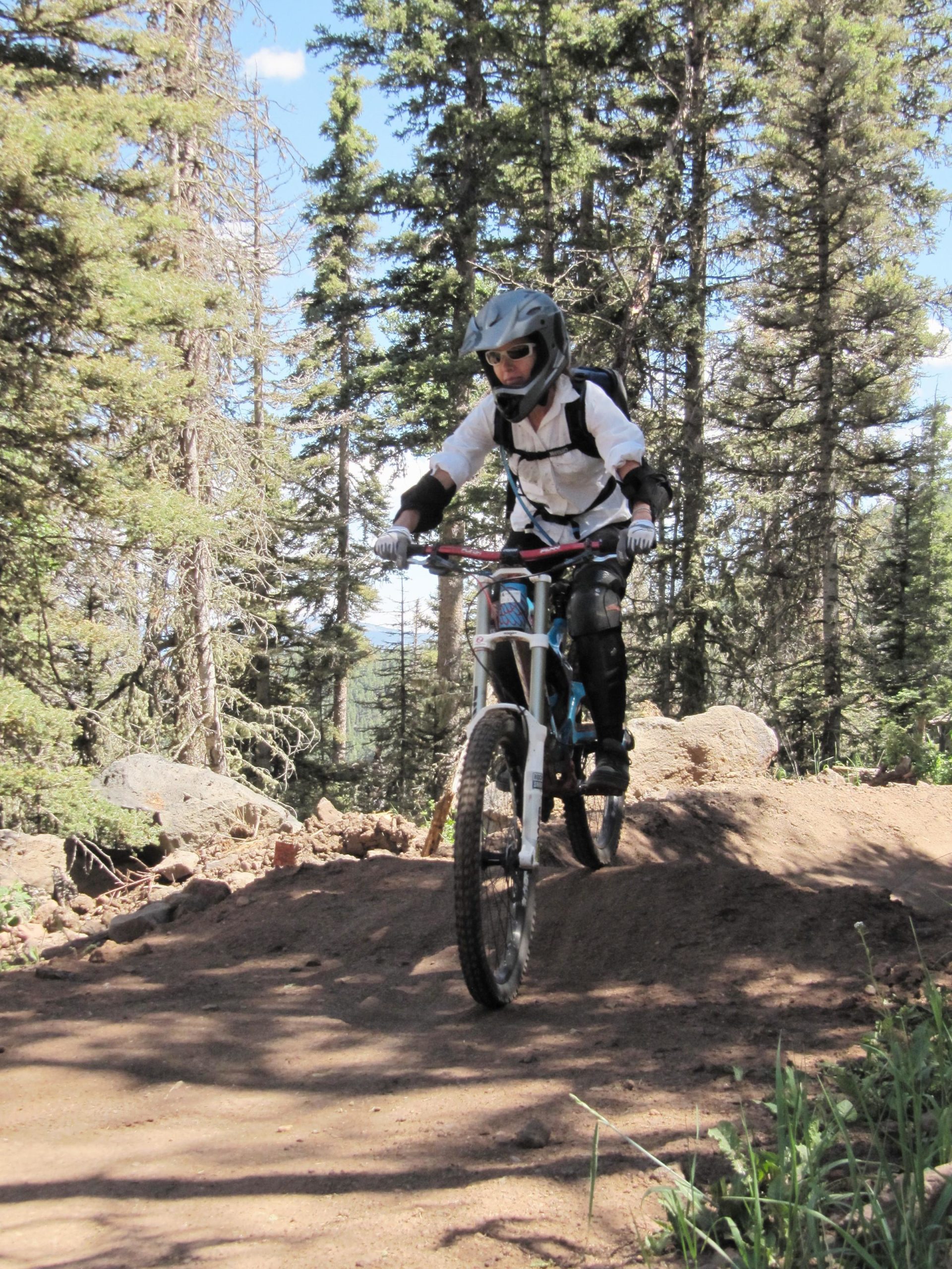 A person riding a mountain bike on a dirt trail in a forested area, wearing a helmet and protective gear. The background features tall trees and a clear blue sky. The rider is navigating over a small dirt mound, demonstrating skill in mountain biking. Angel Fire Bike Park mountain bike trail.
