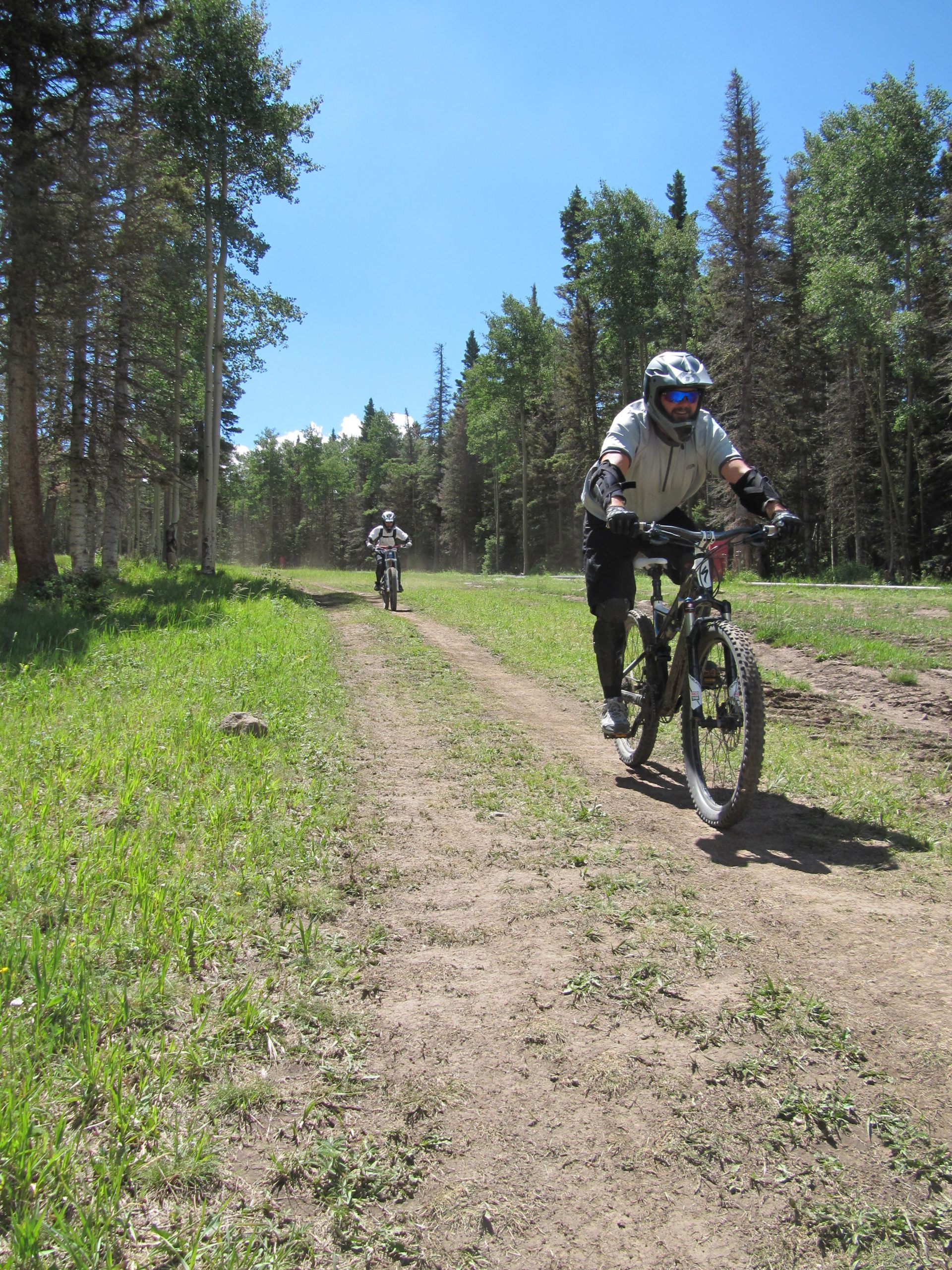 Two mountain bikers ride along a dirt path in a forested area. The sun shines brightly in a clear blue sky, and trees line both sides of the trail, creating a lush green landscape. One rider is closer to the foreground, wearing protective gear and a helmet, while the other is visible further down the path. Angel Fire Bike Park mountain bike trail.