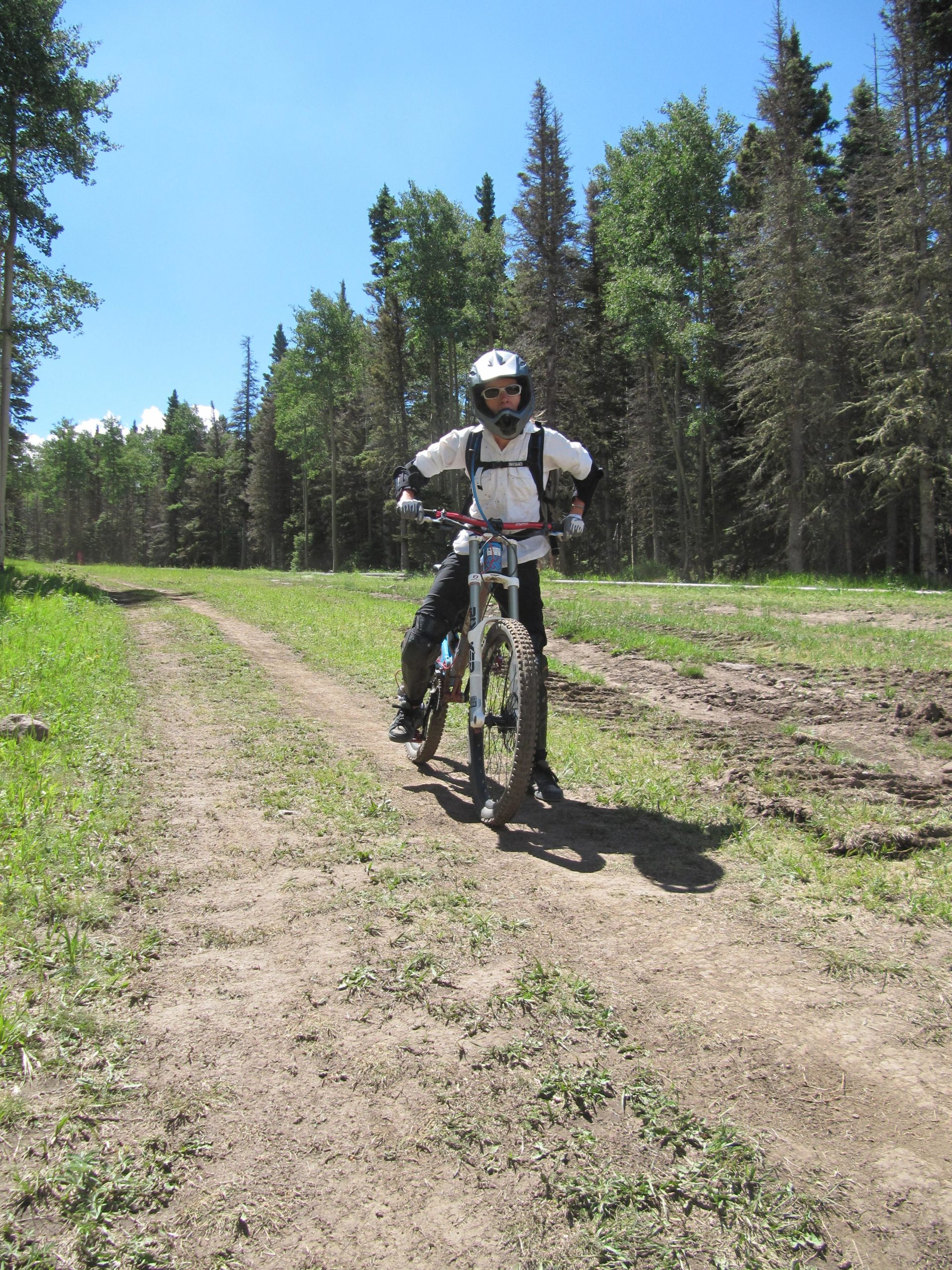 A person wearing a helmet and protective gear is riding a mountain bike on a dirt path surrounded by green grass and tall trees under a clear blue sky. Angel Fire Bike Park mountain bike trail.