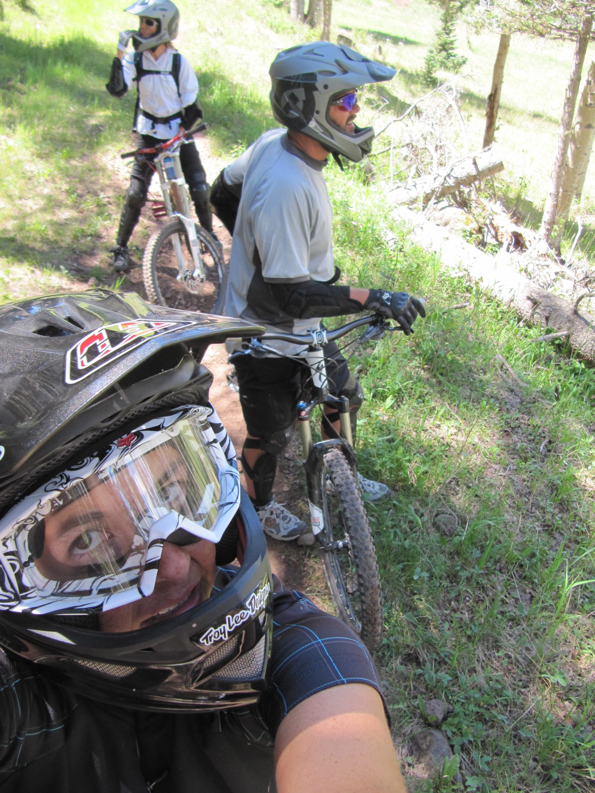 Three individuals dressed in mountain biking gear are on a forested trail. The person in the foreground is taking a selfie, wearing a black helmet with visor and a dark shirt. In the background, two others are seen, one in a white jacket and the other in a gray shirt, both positioned next to their bikes. The surrounding area features green grass and trees, suggesting a sunny day in the woods. Angel Fire Bike Park mountain bike trail.