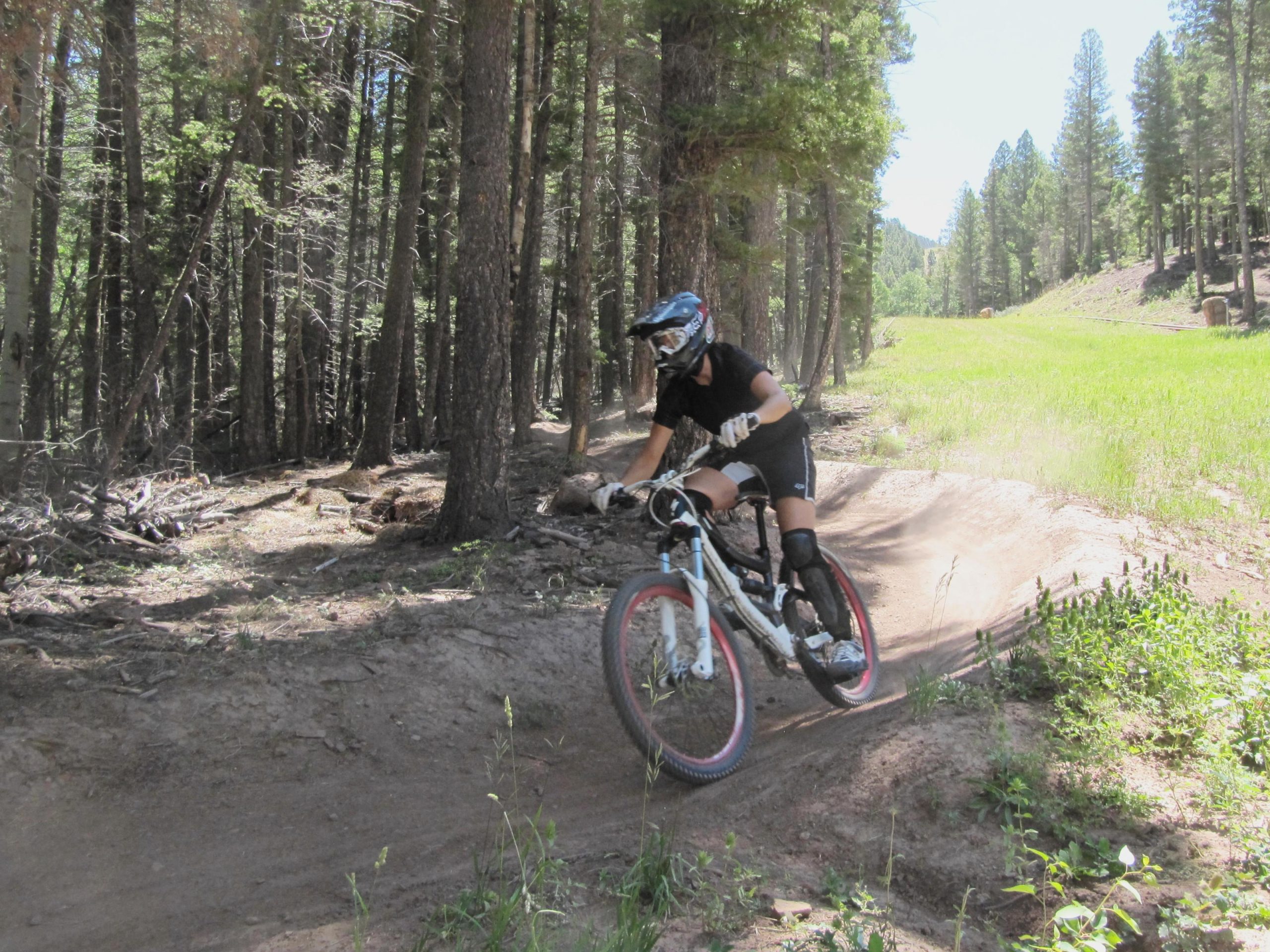 A person riding a mountain bike on a dirt trail in a forested area. The cyclist is wearing protective gear, including a helmet and knee pads, as they navigate a turn. Sunlight filters through the trees, illuminating the green grass and surrounding foliage. Angel Fire Bike Park mountain bike trail.