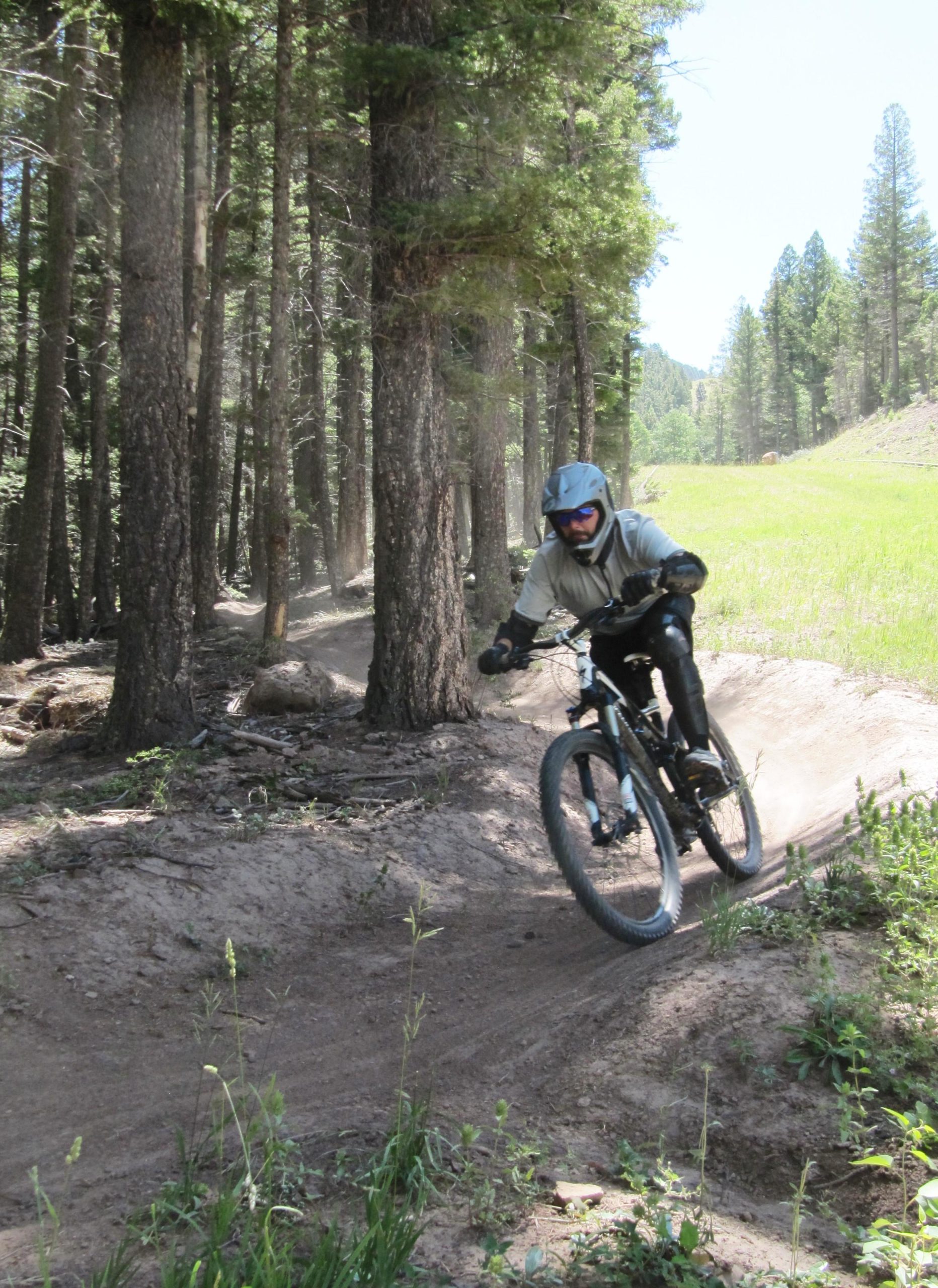 A mountain biker wearing a helmet and protective gear is navigating a dirt path through a forest, surrounded by tall trees. The rider leans into a turn, kicking up dust on the trail, with green grass visible in the background. Angel Fire Bike Park mountain bike trail.