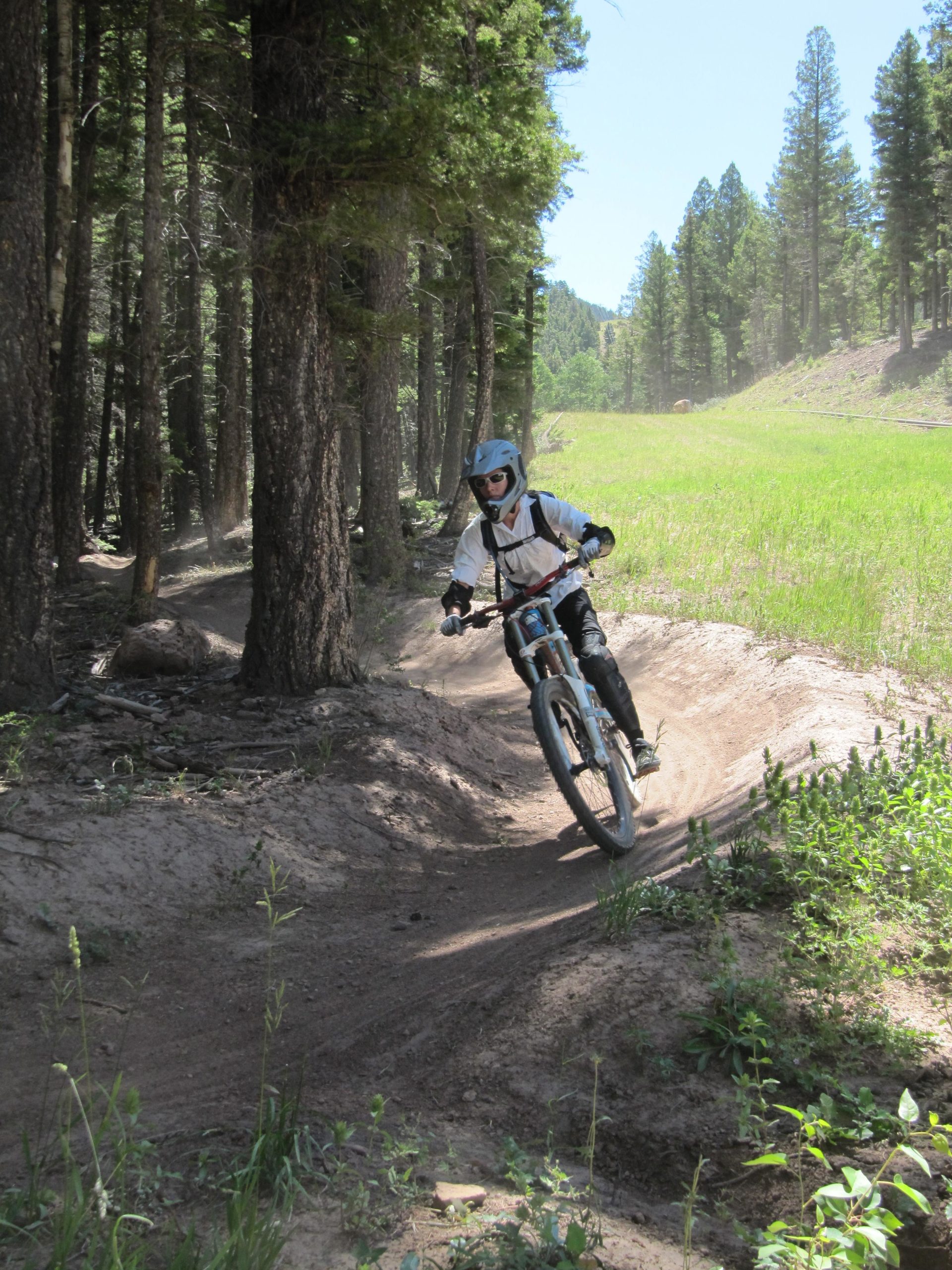 A mountain biker riding along a dirt trail in a forested area, surrounded by tall trees and green grass, with sunlight filtering through the foliage. The rider is wearing a helmet and protective gear while navigating a curve in the path. Angel Fire Bike Park mountain bike trail.