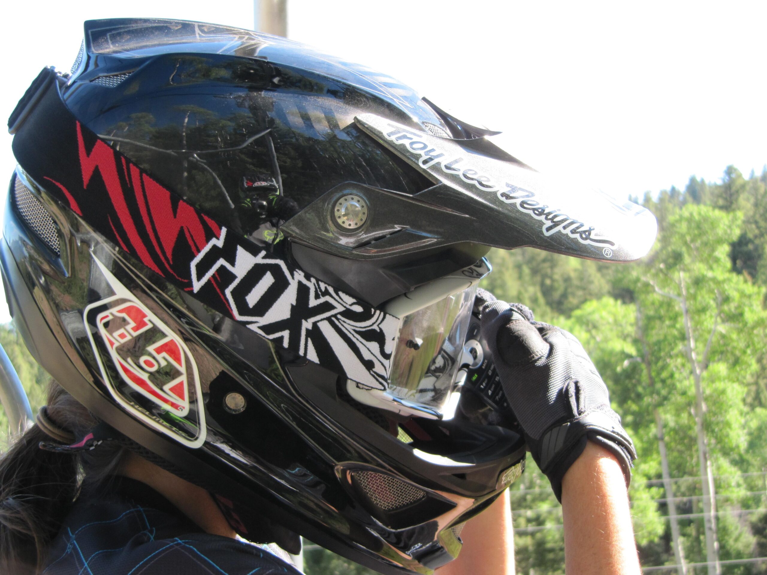A close-up image of a black motocross helmet adorned with red and white graphics, held by a person wearing black gloves. The helmet features logos from Troy Lee Designs and Fox Racing, and the background shows a green, forested landscape under bright sunlight. Angel Fire Bike Park mountain bike trail.