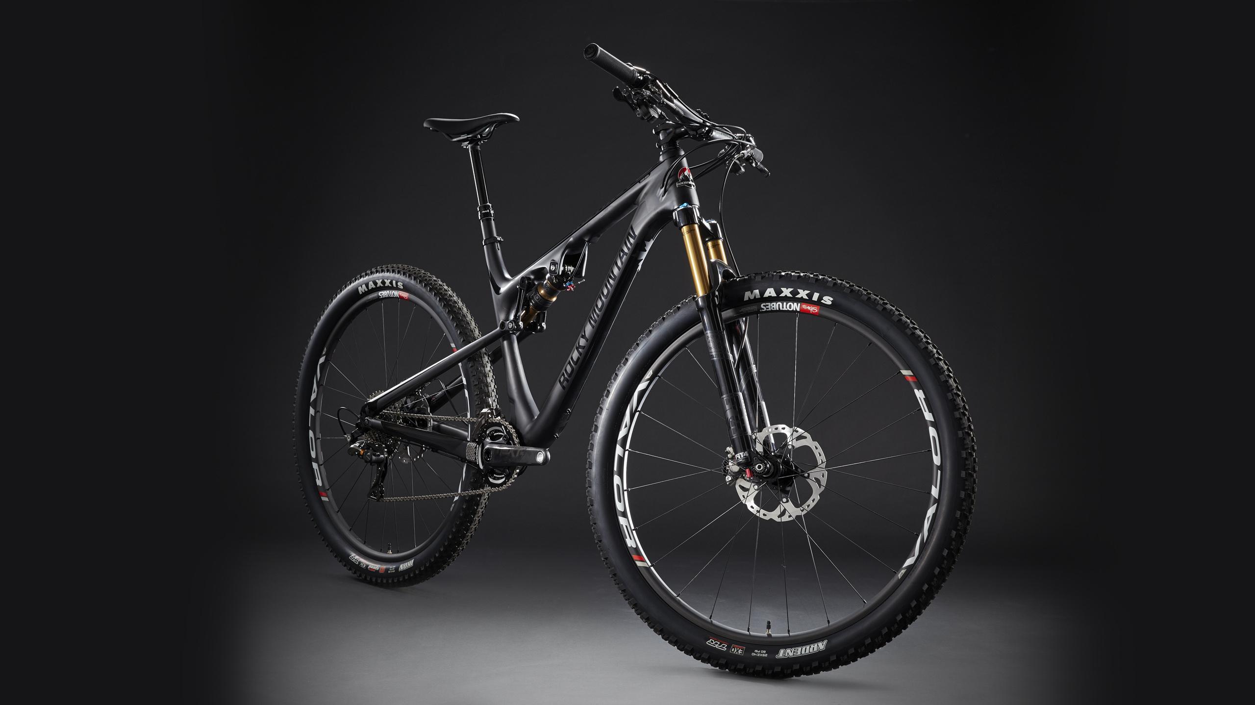 Rocky Mountain Instinct 999 MSL: Image of a modern mountain bike featuring a sleek black frame with gold suspension forks. The bike has wide, knobby tires labeled "Maxxis" and clearly visible disc brakes. The background is dark, emphasizing the bike's design and components.